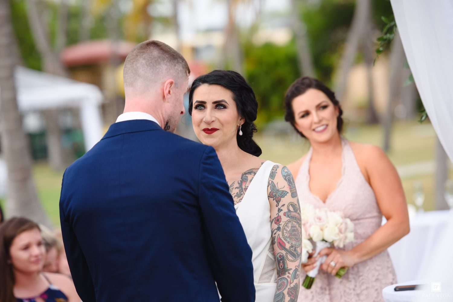 Bride with tattoos exchanges vows with groom in navy suit as smiling bridesmaid holds bouquet at outdoor wedding.