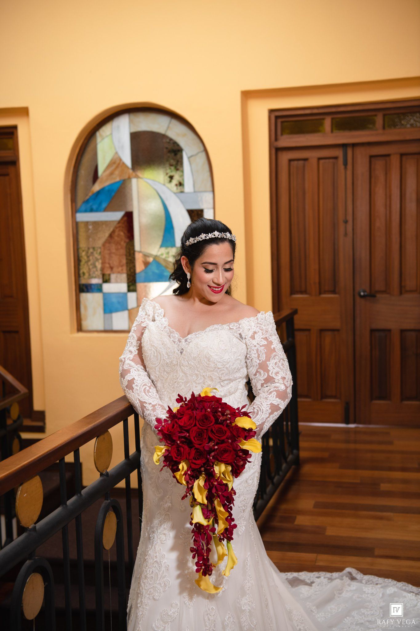 Bride in lace off-shoulder gown holding red and yellow cascading bouquet on staircase near stained glass window.