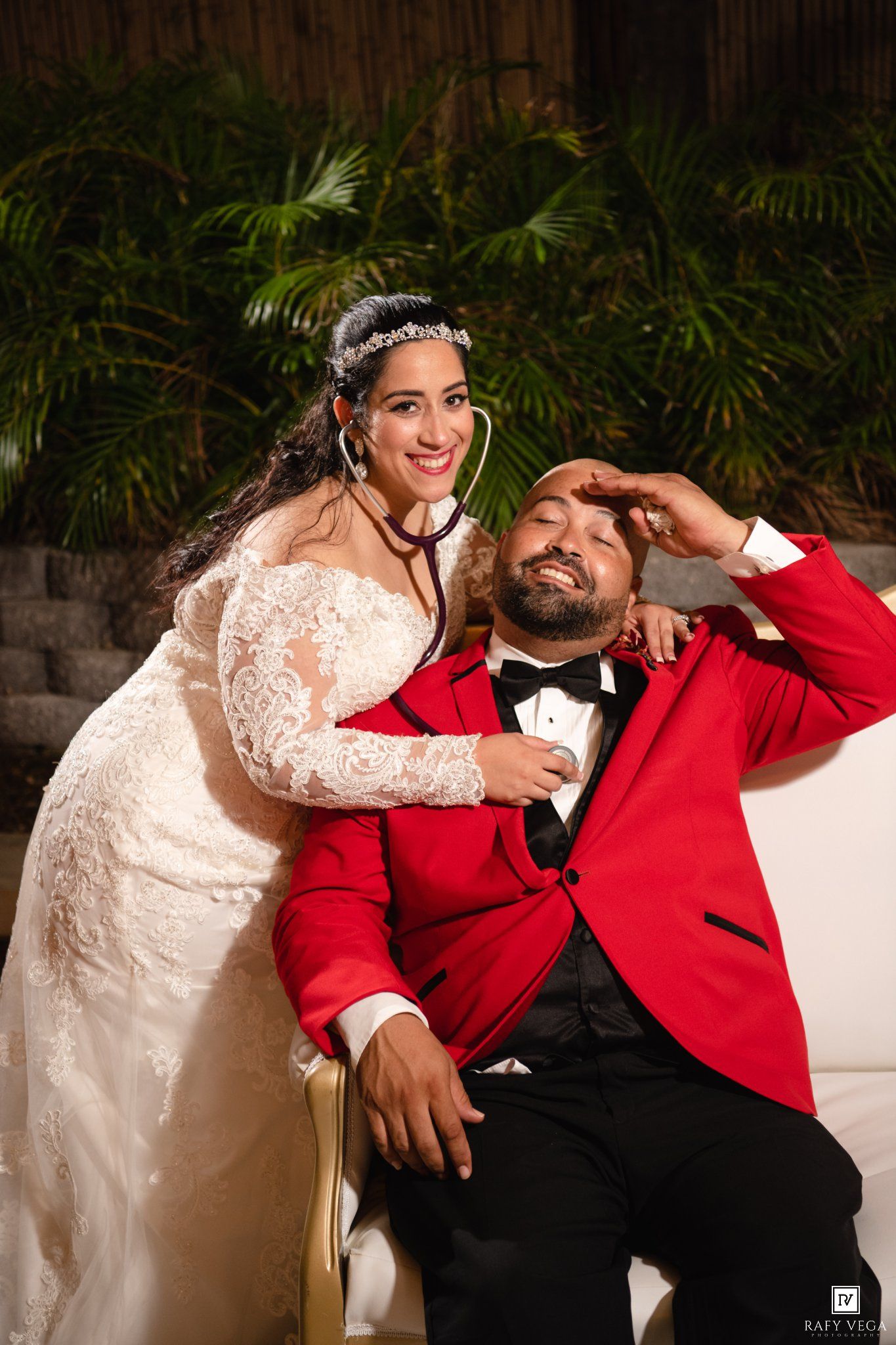 Bride in lace gown uses stethoscope on groom in red tuxedo jacket at elegant outdoor wedding photo.