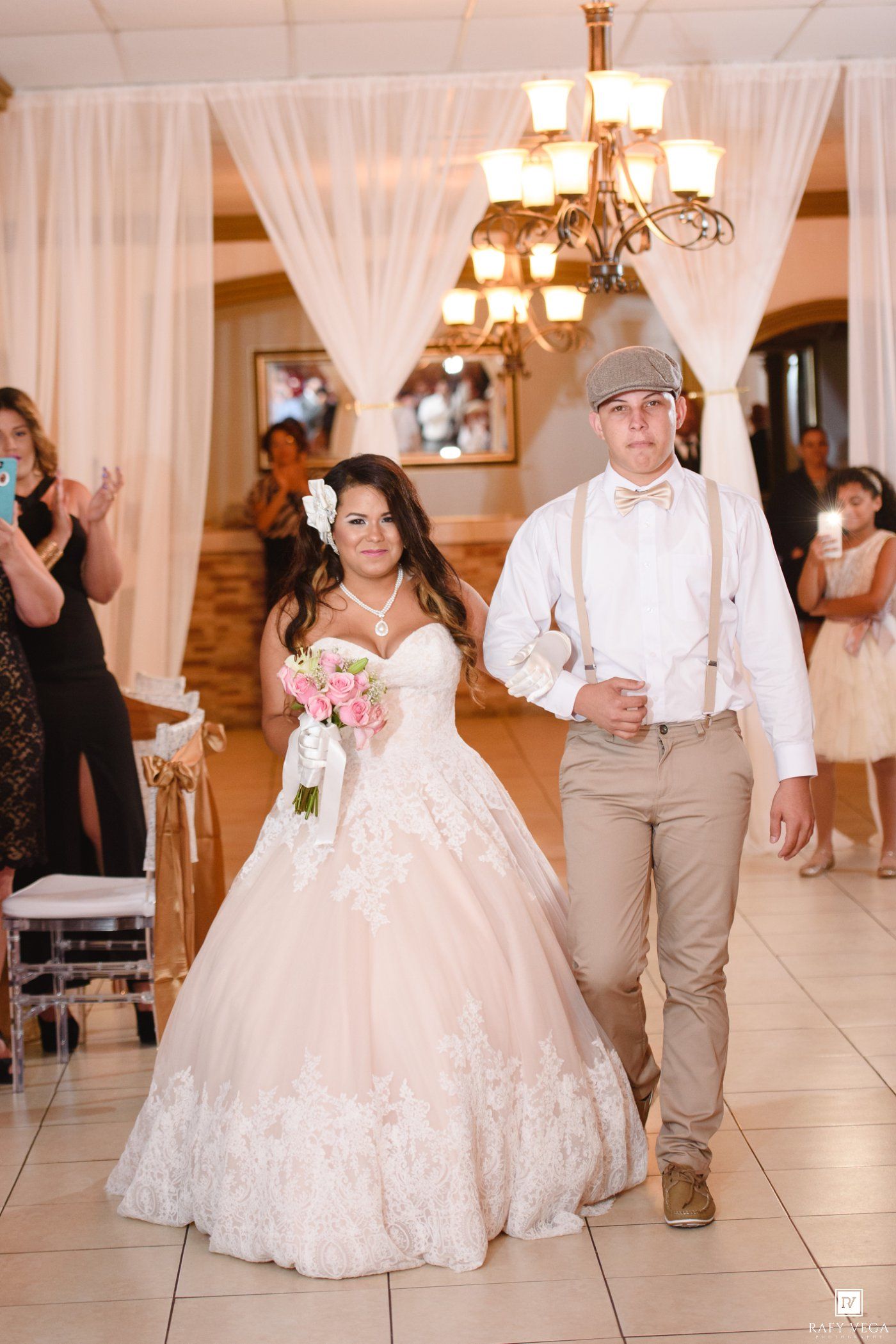 Bride in blush ballgown and groom in suspenders make grand reception entrance under chandelier.