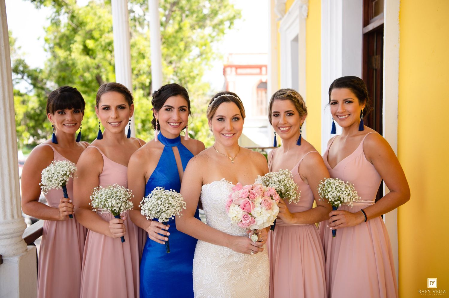 Bride in white lace gown poses with five bridesmaids in blush pink dresses holding baby's breath bouquets outdoors.