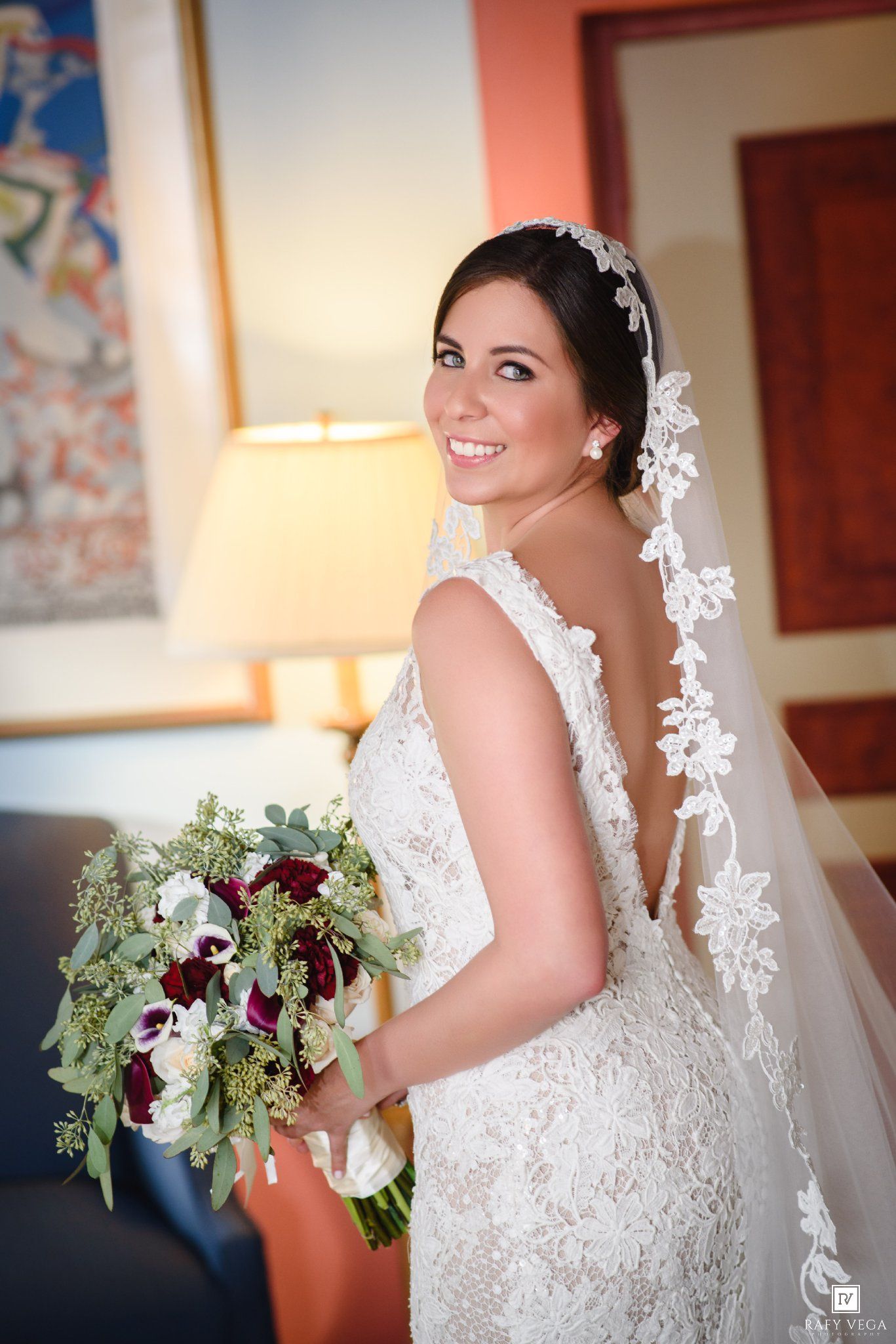 Bride in backless lace gown holds burgundy bouquet, lace veil trailing down her back.