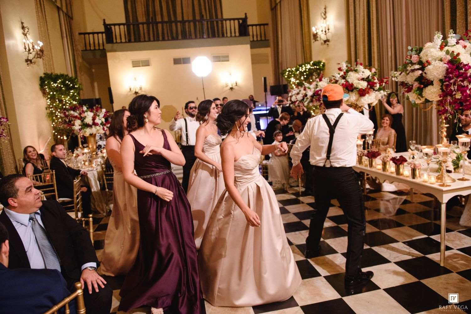 Bridesmaids in purple and gold gowns dance on a black and white checkered floor at an elegant wedding reception.