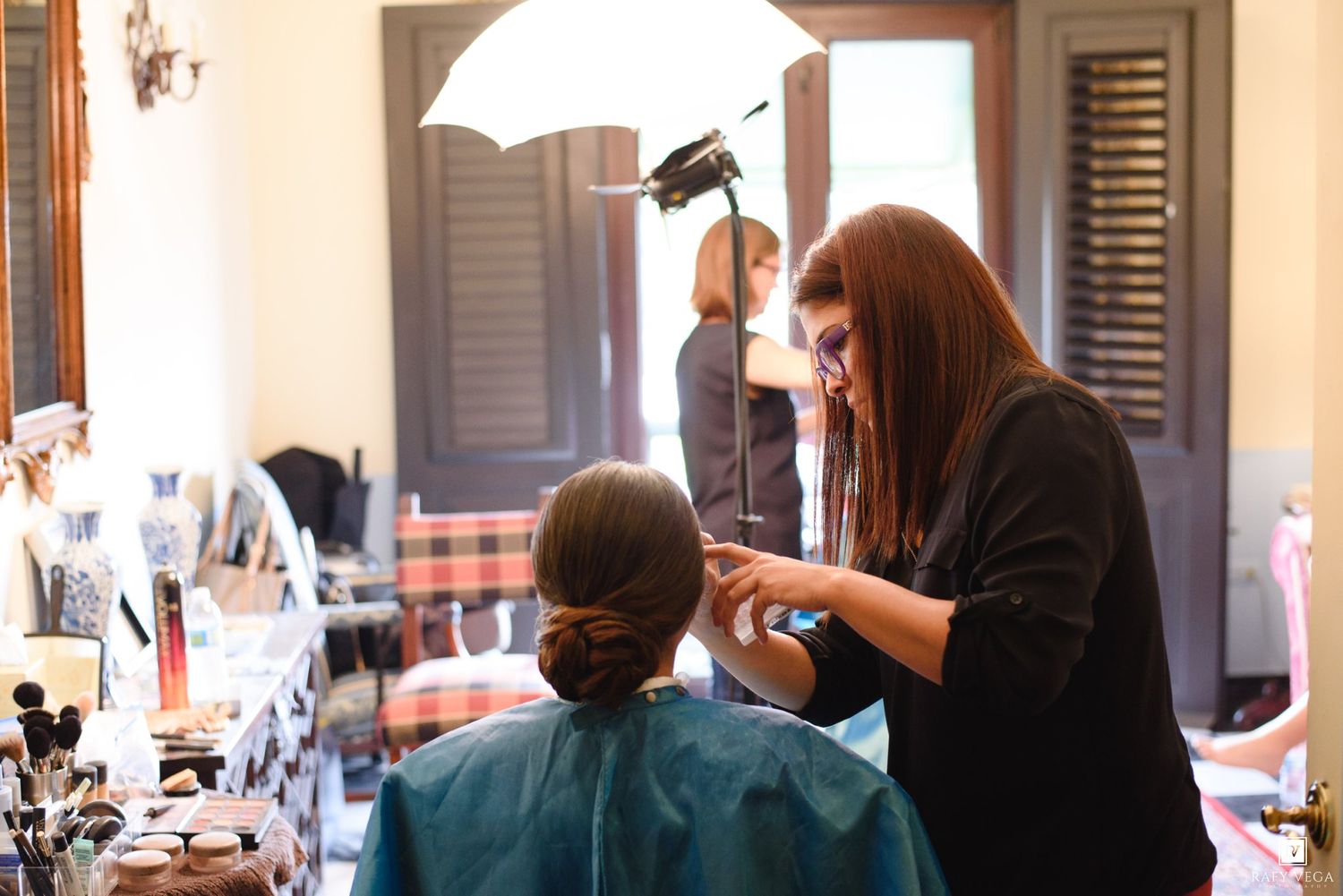 A hairstylist applying finishing touches to a client's updo in a well-lit salon with makeup products nearby.