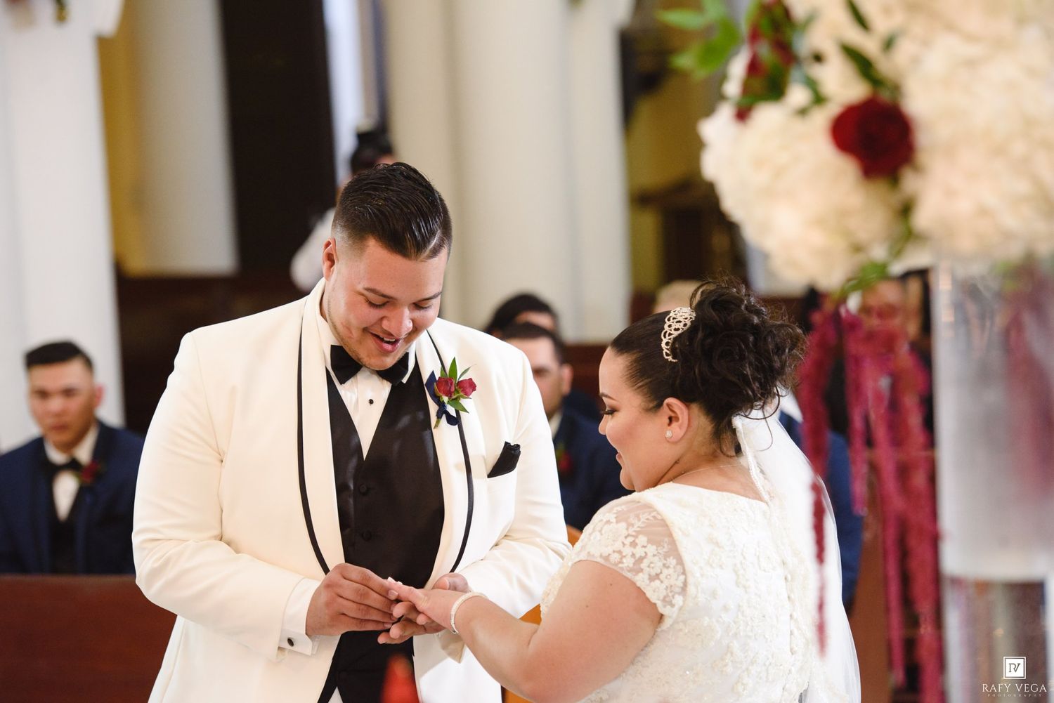 Groom in white tuxedo places wedding ring on bride's finger during elegant church ceremony with floral arrangements.