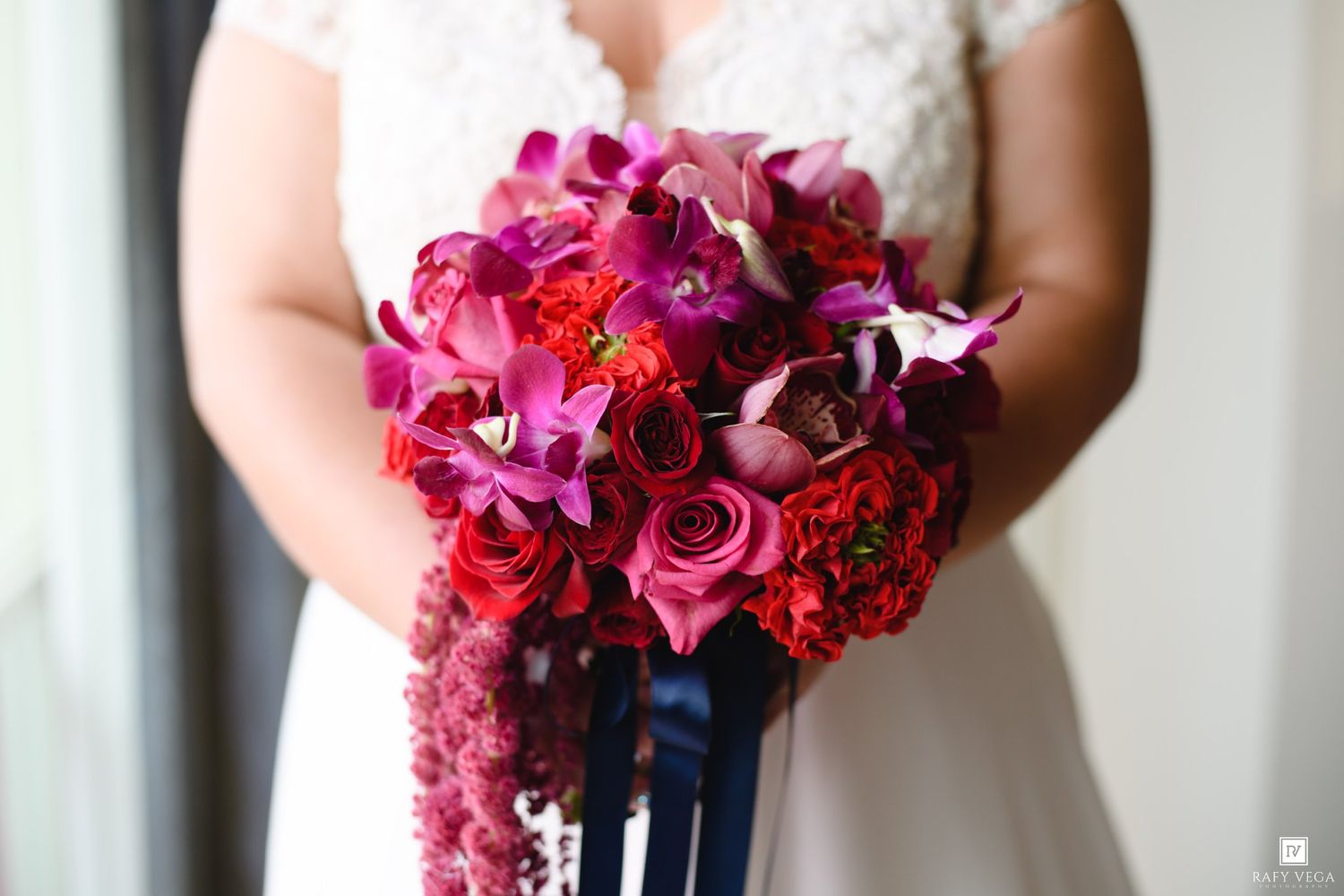 Bride holding a lush bridal bouquet of red roses, purple orchids, and magenta blooms with navy ribbon streamers.