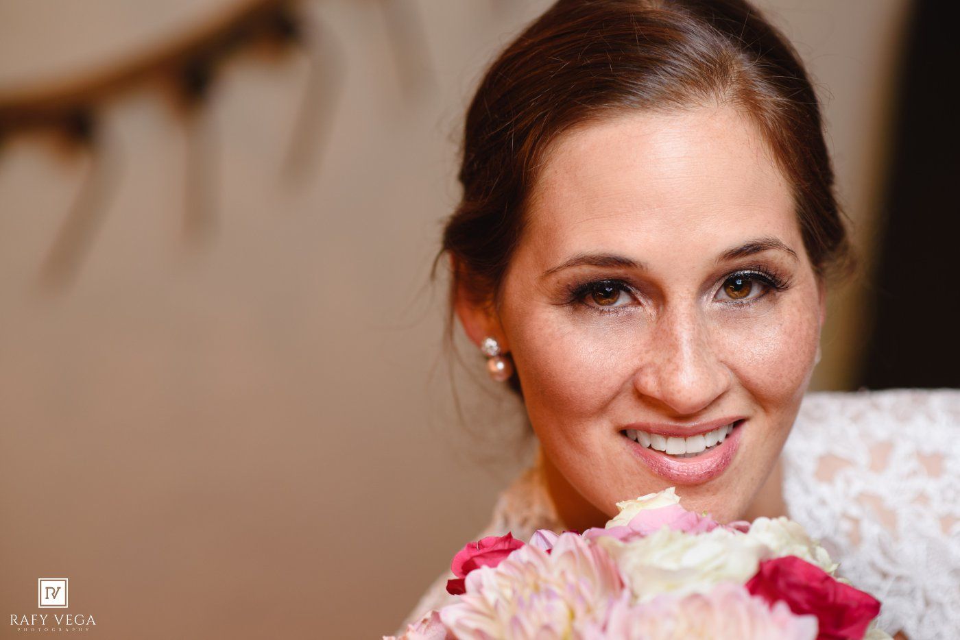 Smiling bride holding pink and white bouquet, wearing pearl earrings and lace dress on her wedding day.