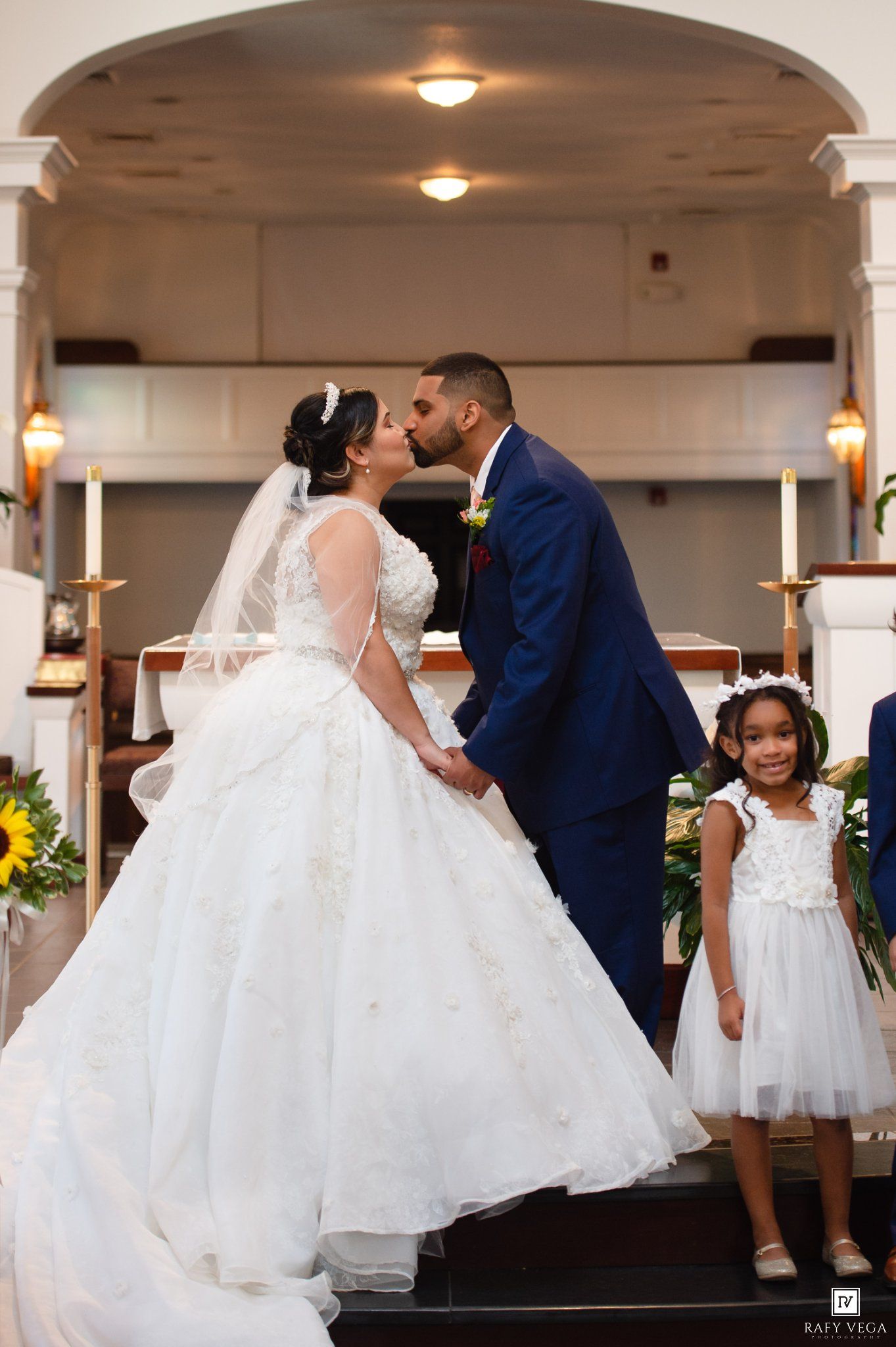 Bride and groom share first kiss at church altar with flower girl beside them during wedding ceremony.