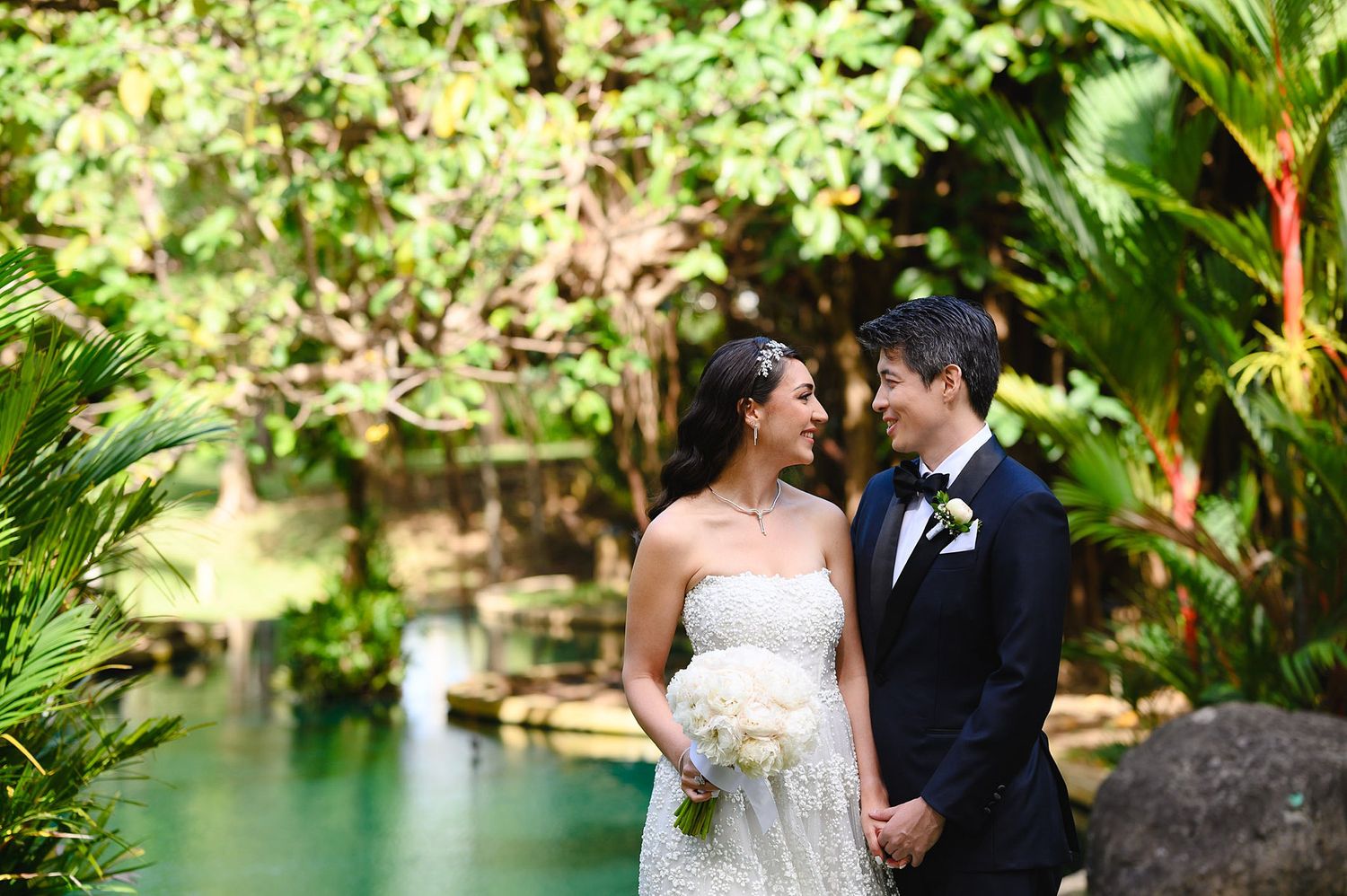 Bride and groom share a romantic moment in lush tropical garden with turquoise water backdrop on their wedding day.