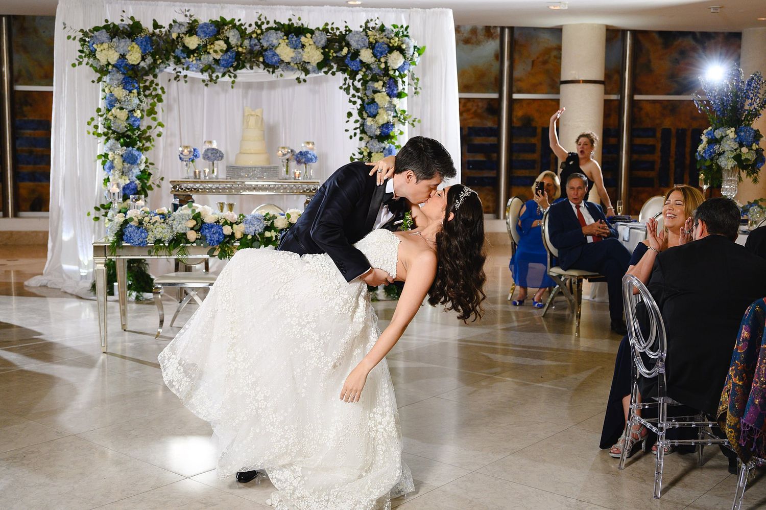 Bride and groom share romantic first dance dip at elegant wedding reception with blue floral arch and wedding cake backdrop.