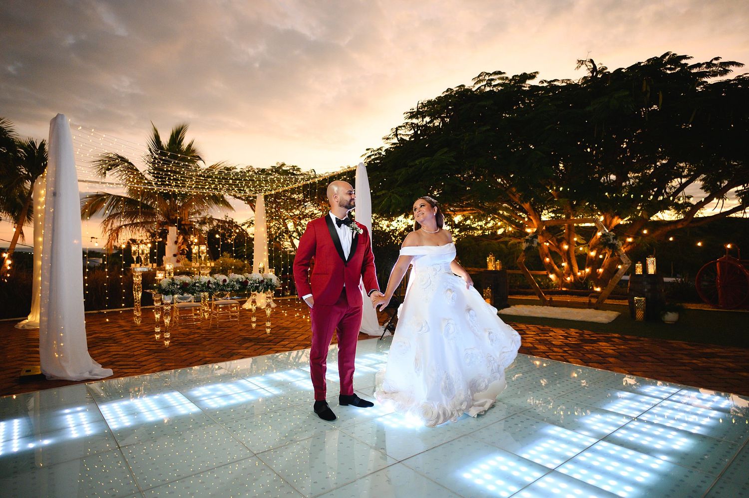 Bride and groom hold hands on illuminated dance floor at elegant outdoor wedding venue with twinkling lights at sunset.