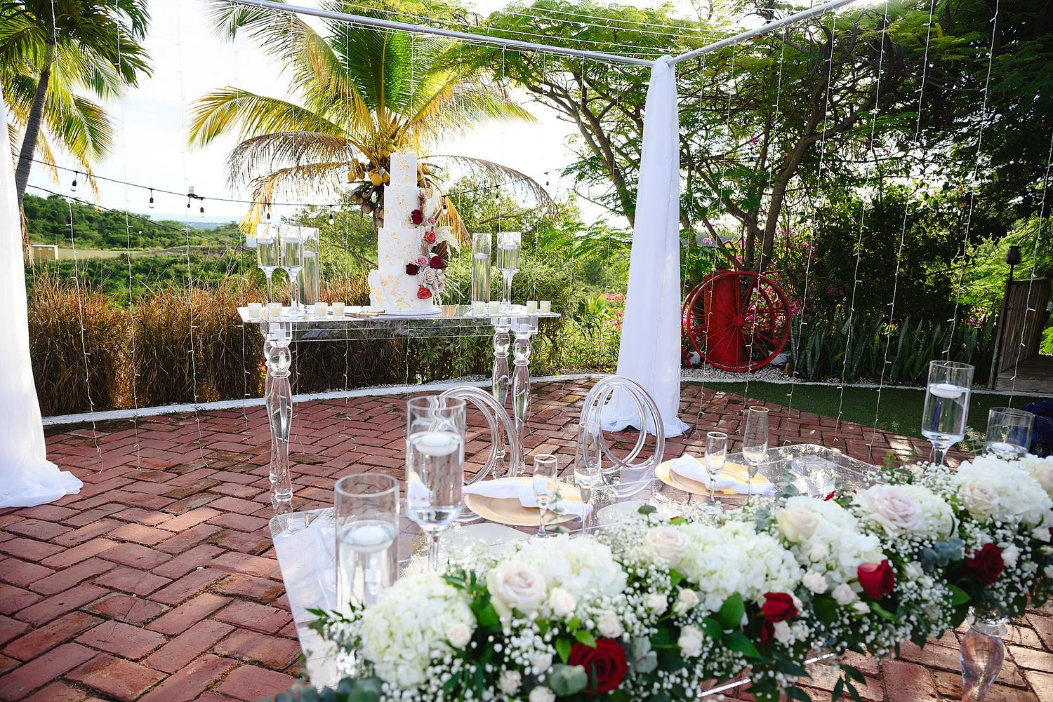 Elegant outdoor wedding reception setup with white floral arrangements, candles, and tropical greenery on a brick patio.
