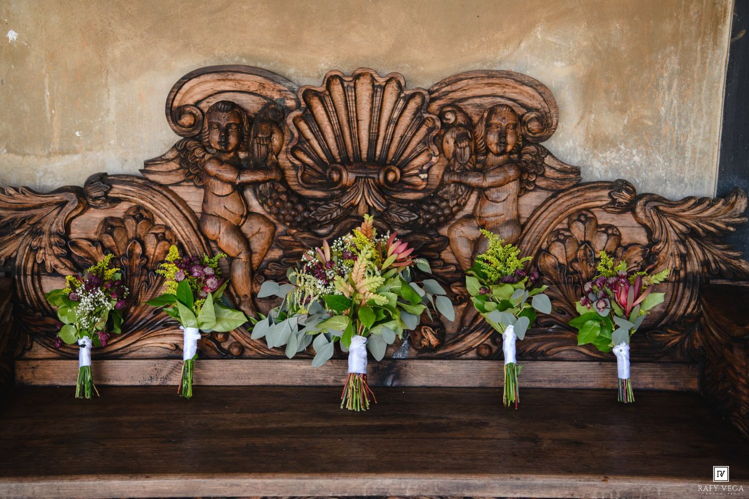 Four colorful wedding bouquets resting on an ornate carved wooden bench with cherub details.