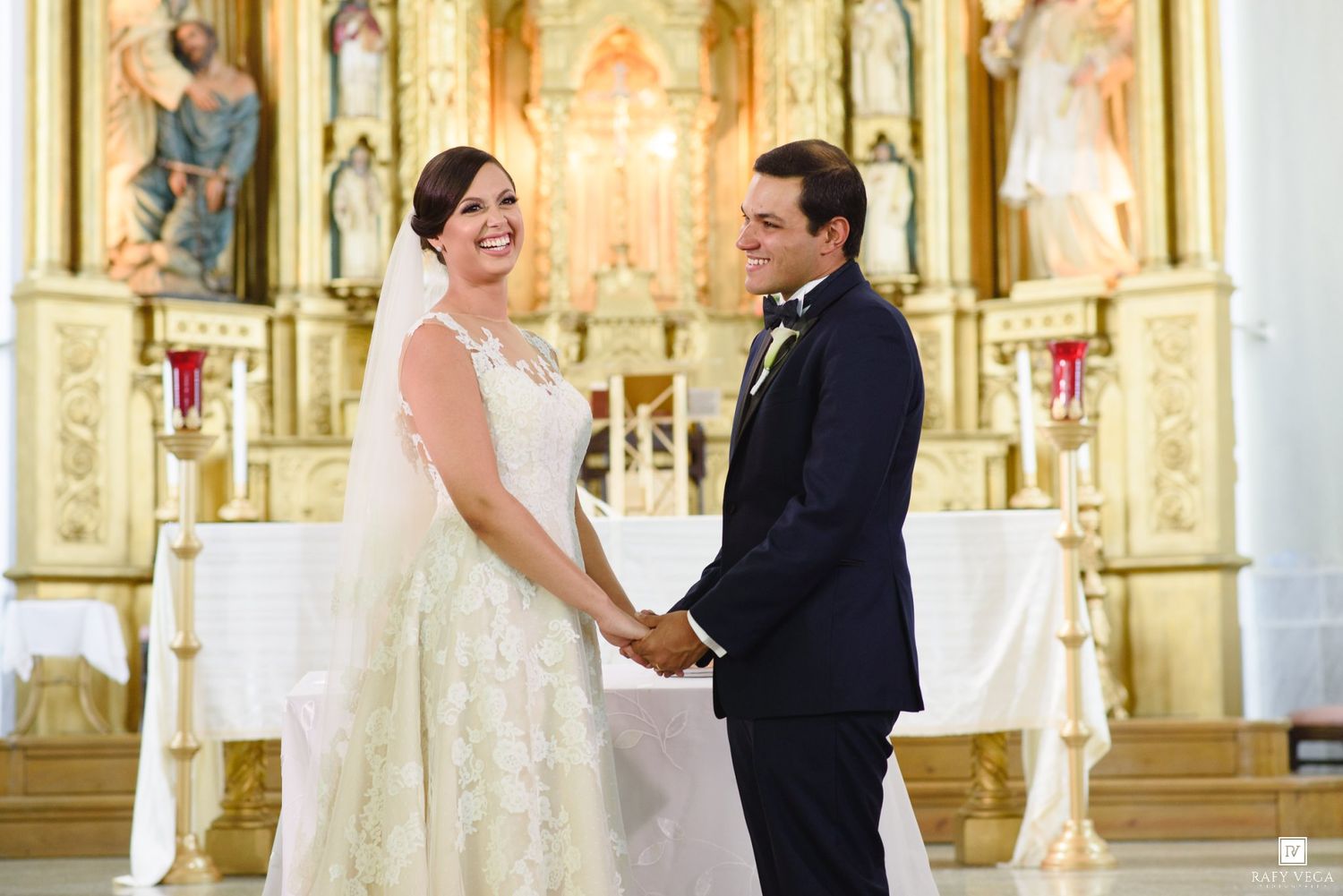 Bride and groom holding hands at church altar during wedding ceremony in white gown and navy suit.