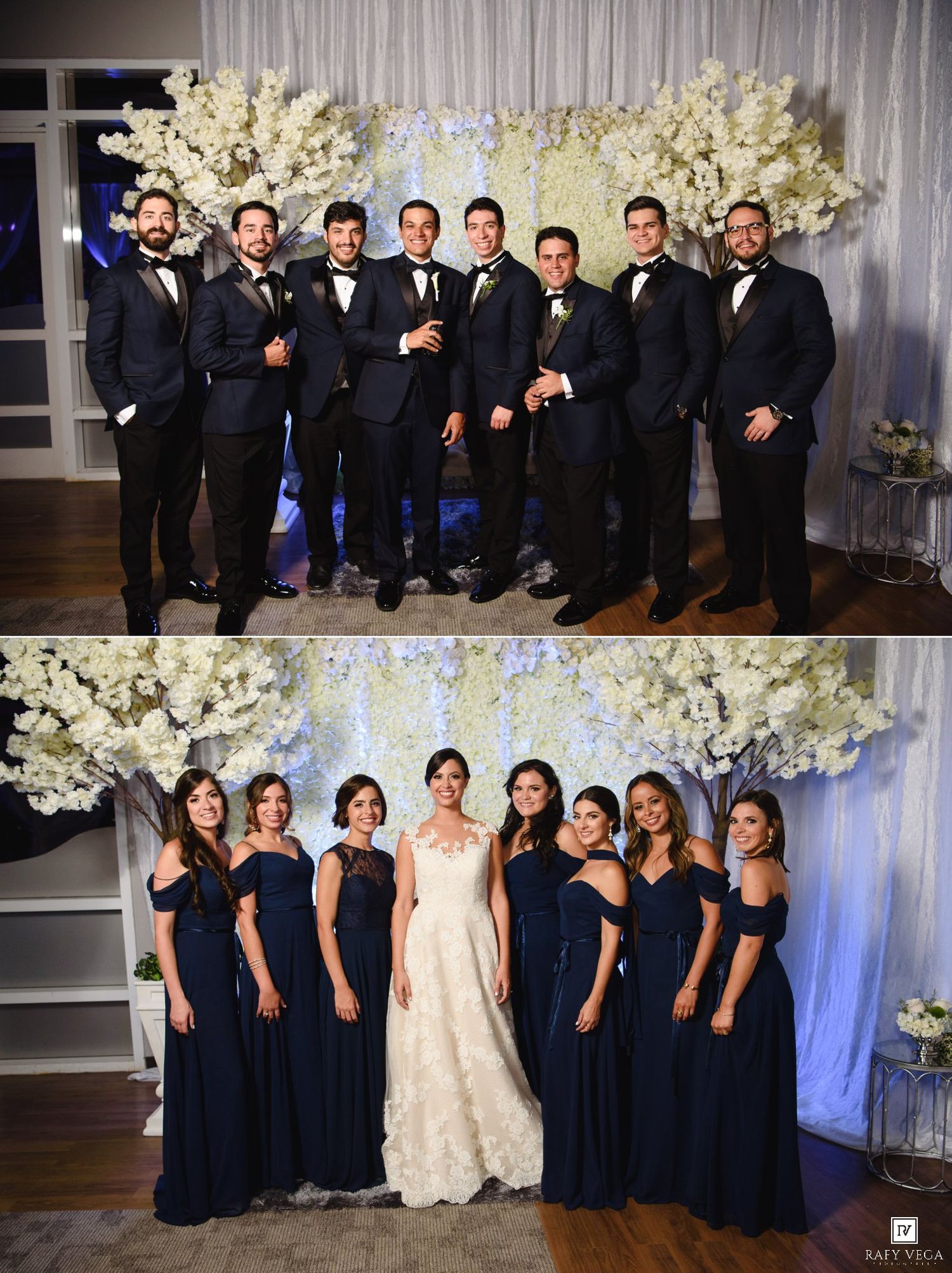 Wedding party in navy attire poses before white floral backdrop, groomsmen top, bridesmaids with bride bottom.