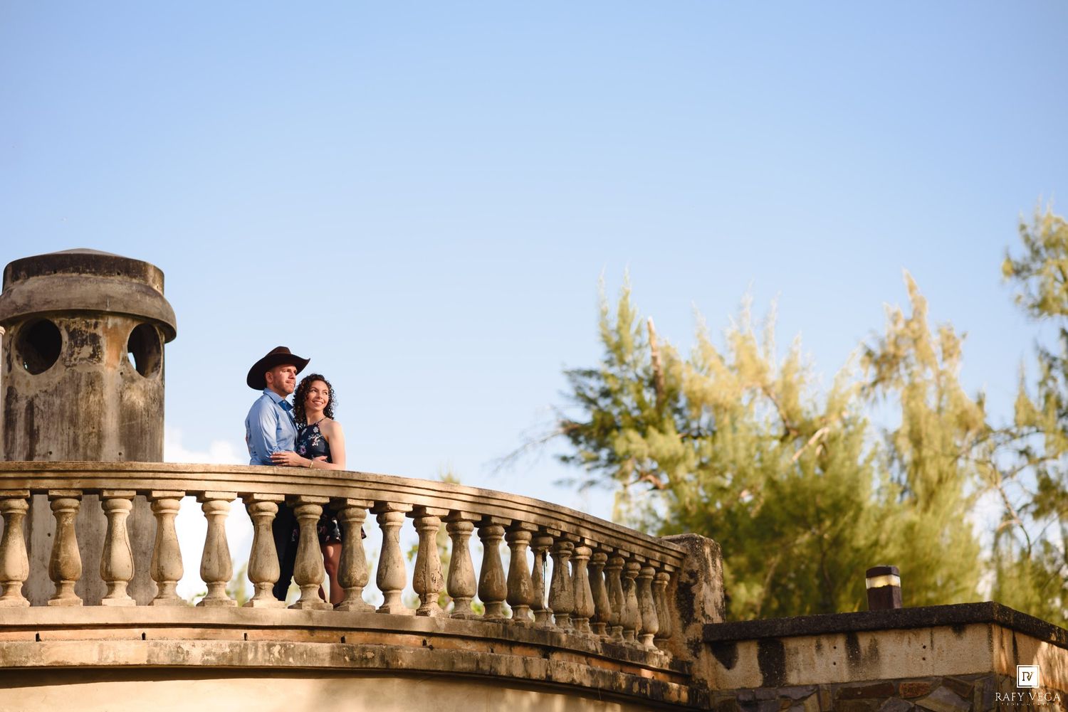 Couple standing on ornate stone balcony balustrade surrounded by tropical trees against clear blue sky.