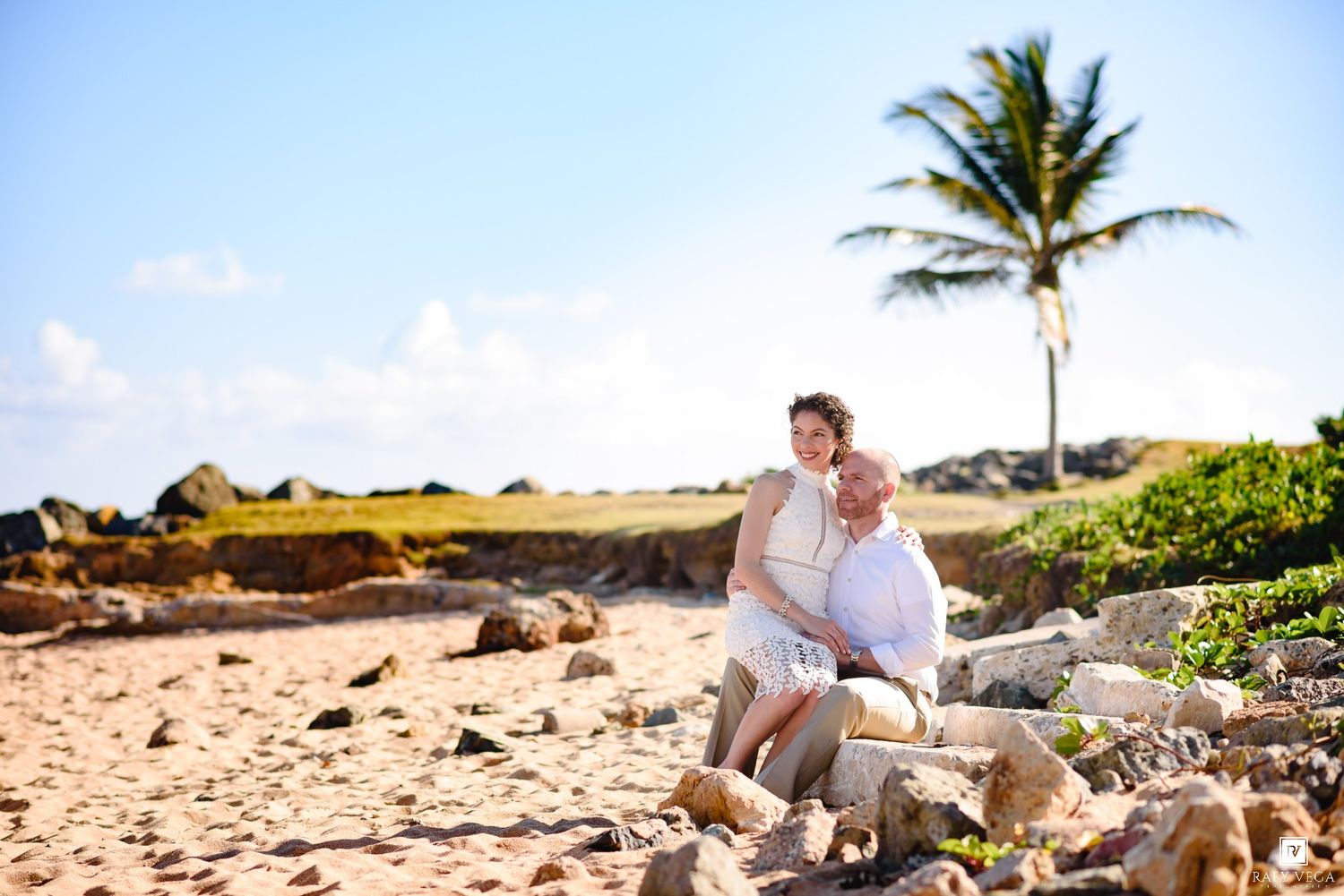 Couple embracing on rocky beach with palm tree and blue sky in tropical destination wedding photo.