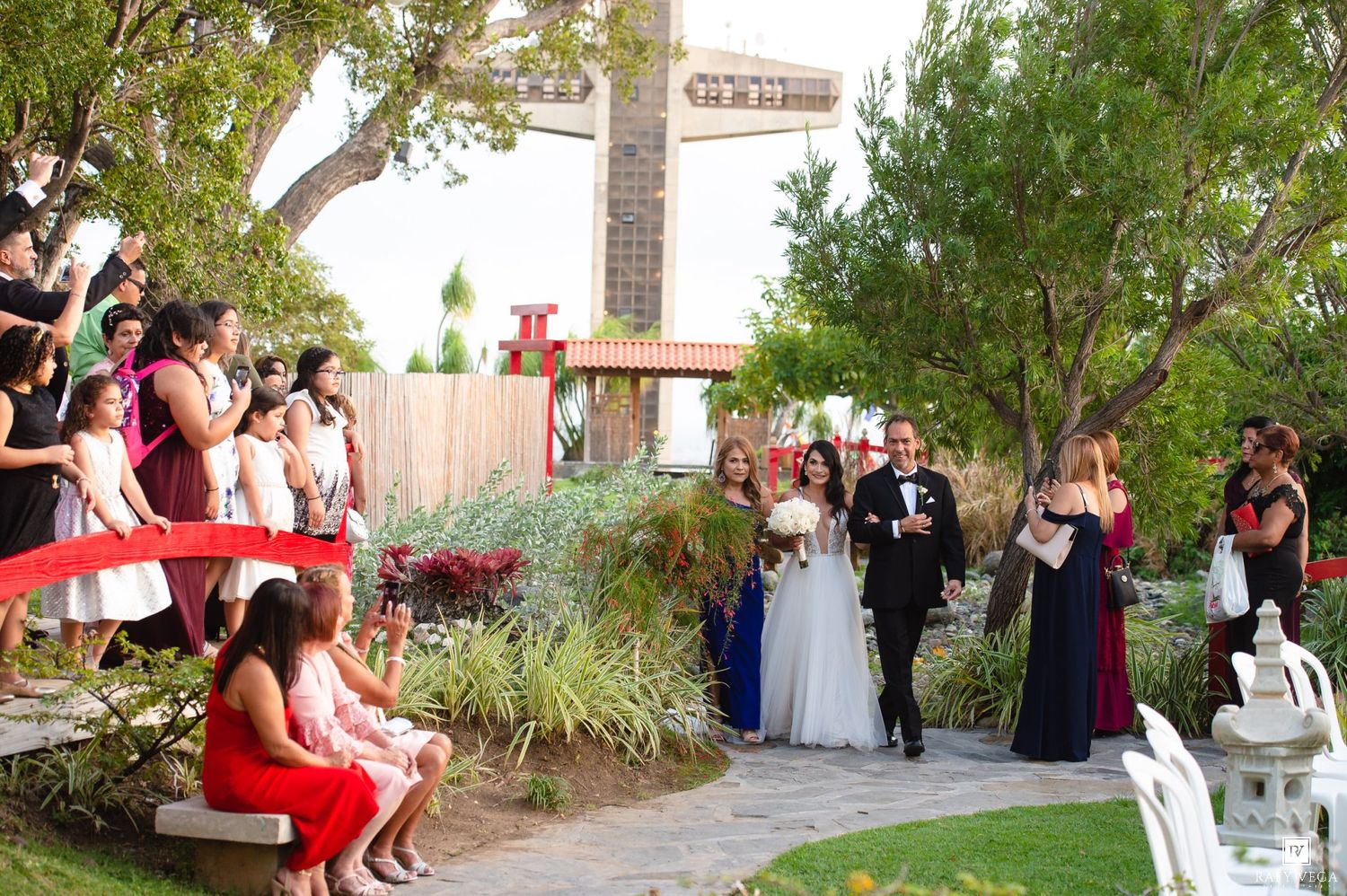Outdoor wedding ceremony on a garden path with bride, groom, and guests in formal attire on a sunny day.