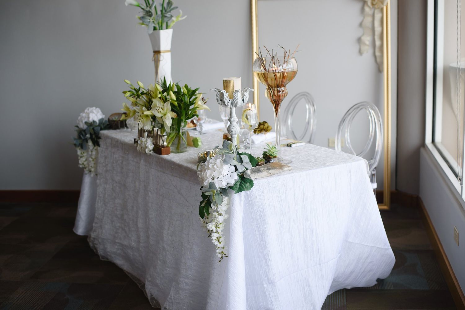 White lace sweetheart table adorned with white floral arrangements, gold candelabra, and clear ghost chairs.