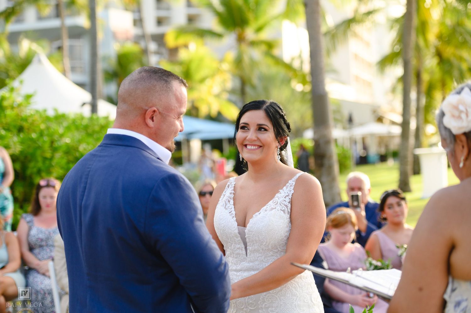 Bride smiling at groom during outdoor beach wedding ceremony with palm trees and guests in background.