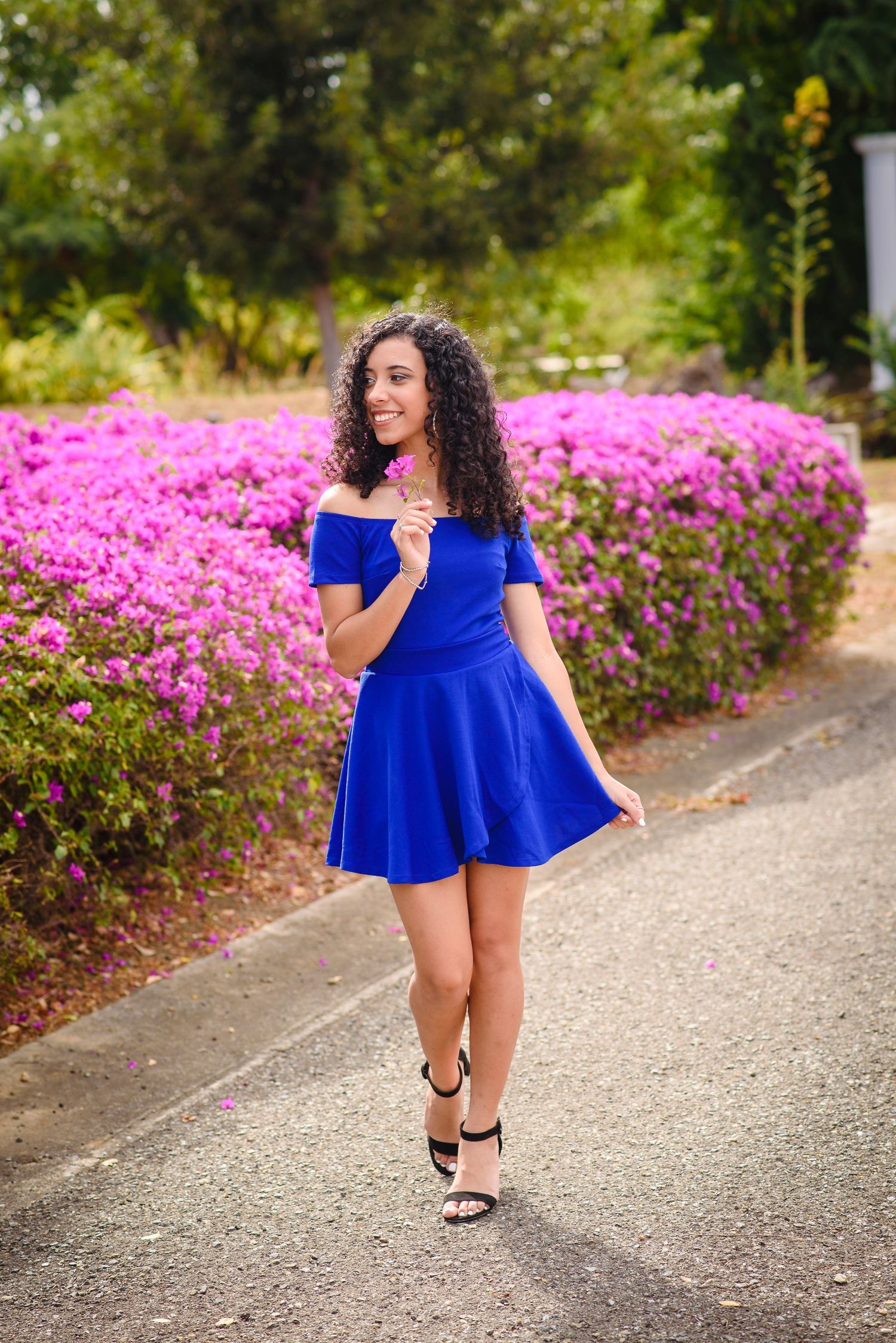 Young woman in blue off-shoulder dress walks past vibrant pink flower bushes, smiling outdoors.