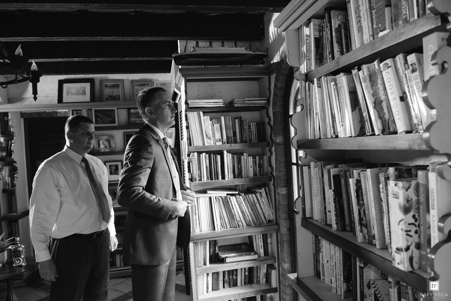 Two men browsing shelves of books in a cozy, well-stocked library room, captured in black and white.