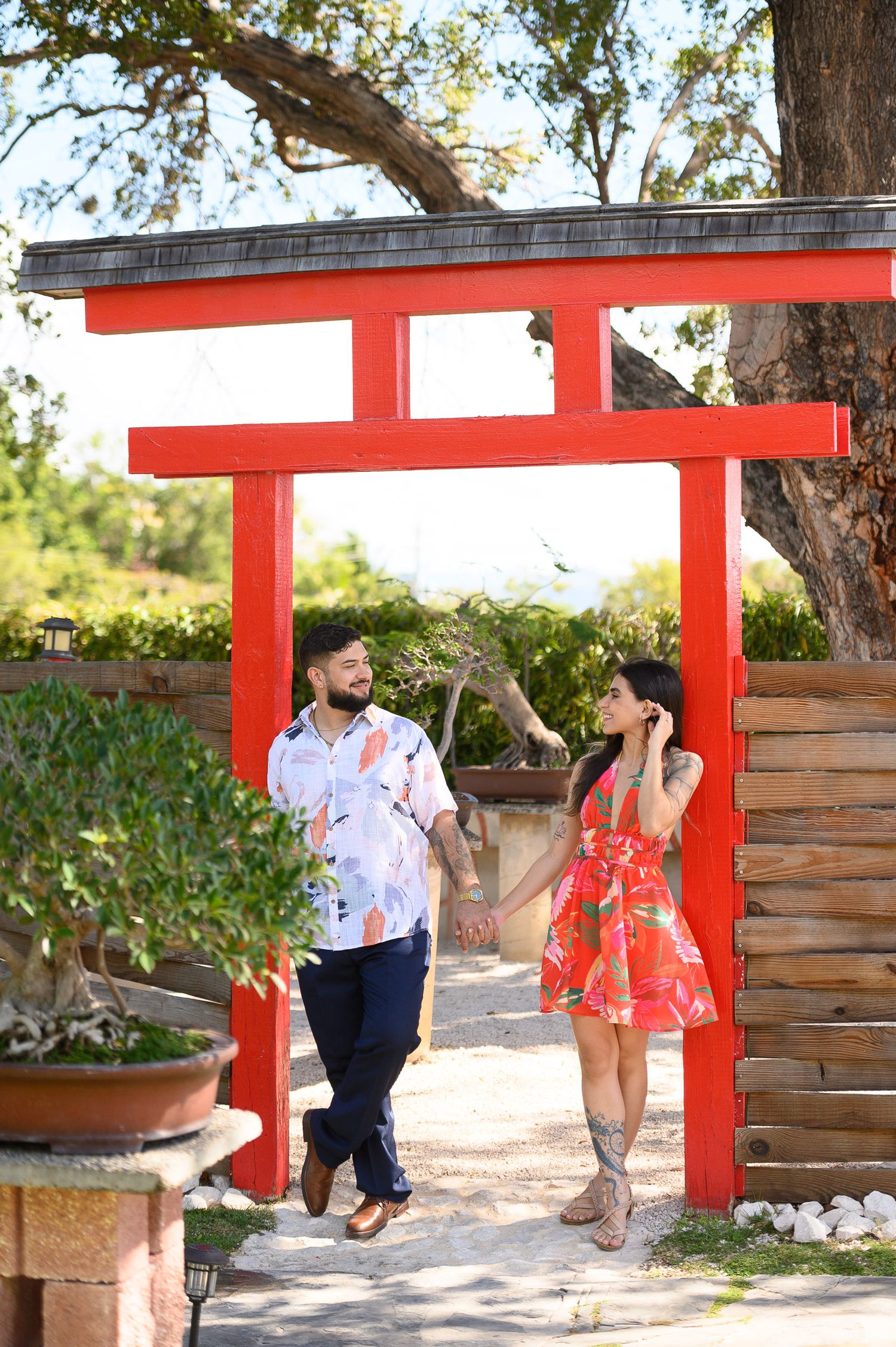 Couple holding hands walking under a red torii gate surrounded by lush greenery, wearing colorful floral outfits.
