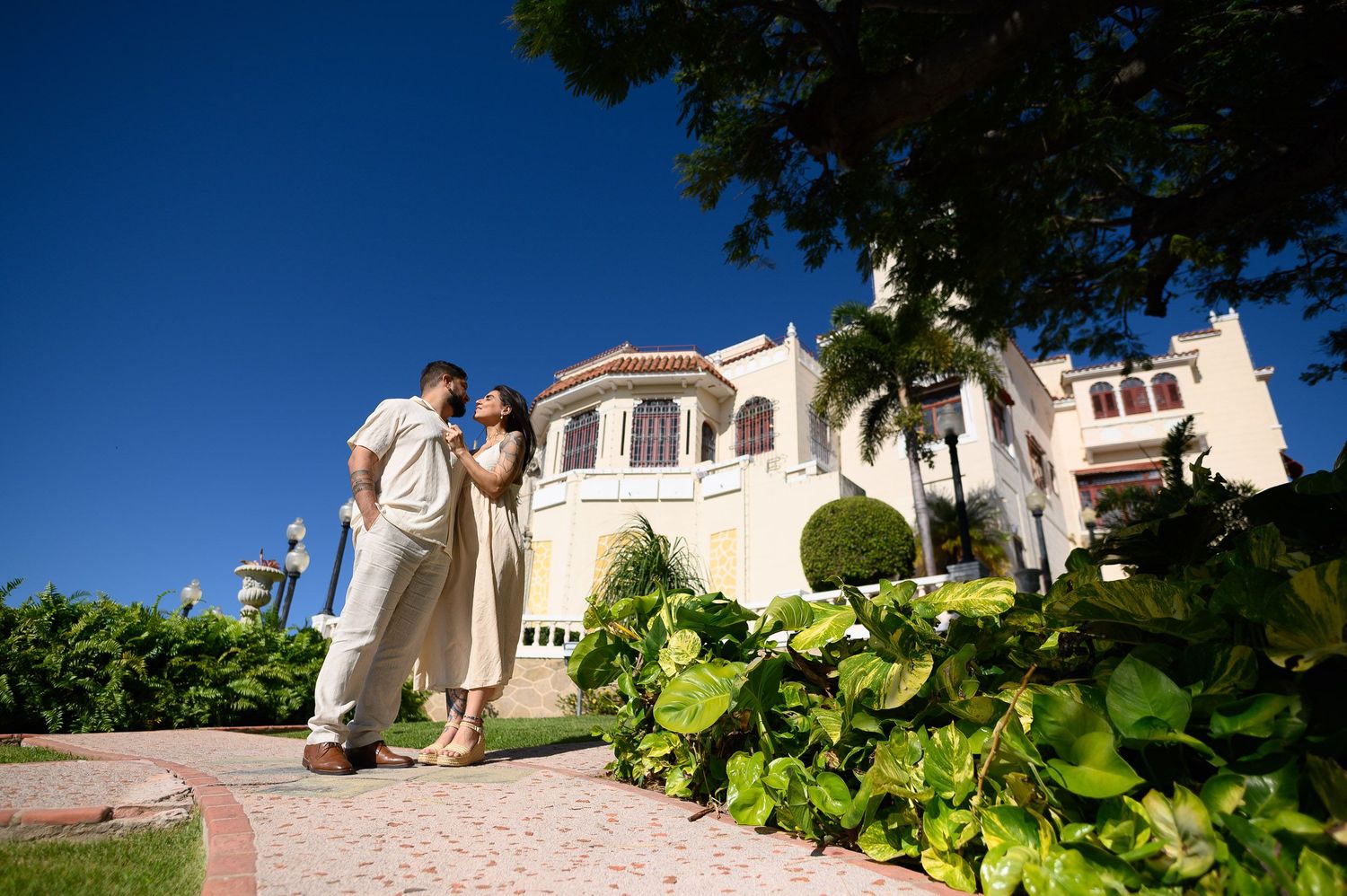 Couple sharing a romantic moment outside a grand tropical villa with lush greenery and blue sky.