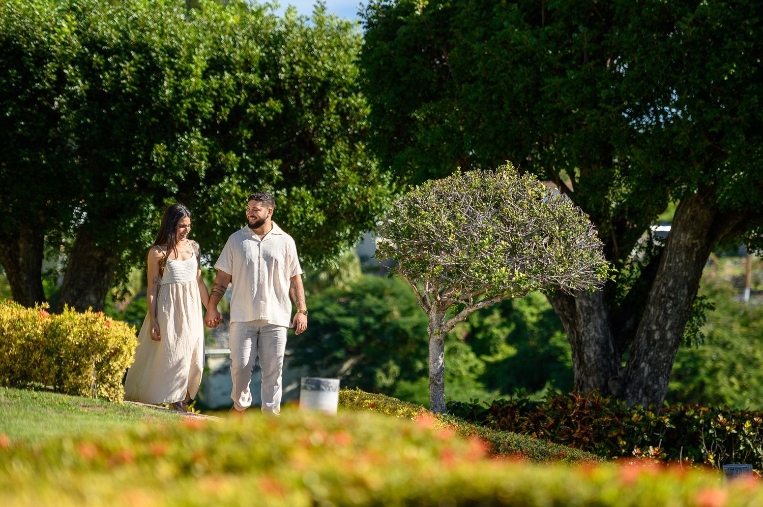 Couple in cream outfits strolling through vibrant green tropical garden on sunny day.