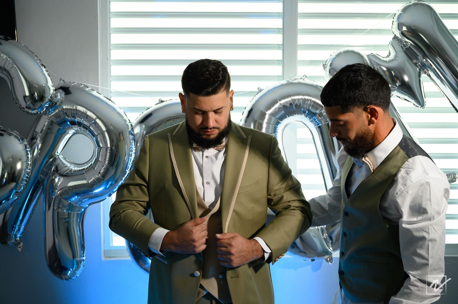 Groom in an olive green suit adjusts his jacket while a groomsman in a vest helps him, with silver balloons nearby.