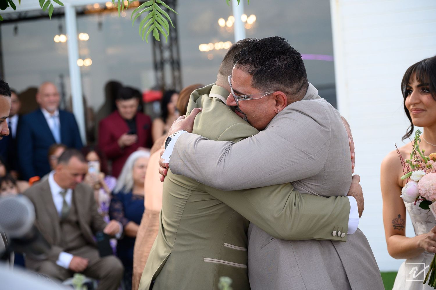 Two men in tan suits share an emotional embrace at an outdoor wedding ceremony while seated guests and a bride holding a bouquet look on.