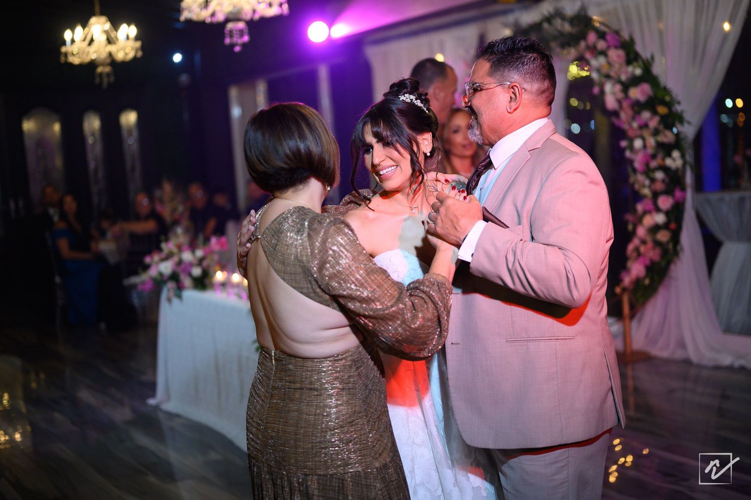A bride in a white gown dances with a man in a beige suit and a woman in a sparkling gold dress at an elegant wedding reception.