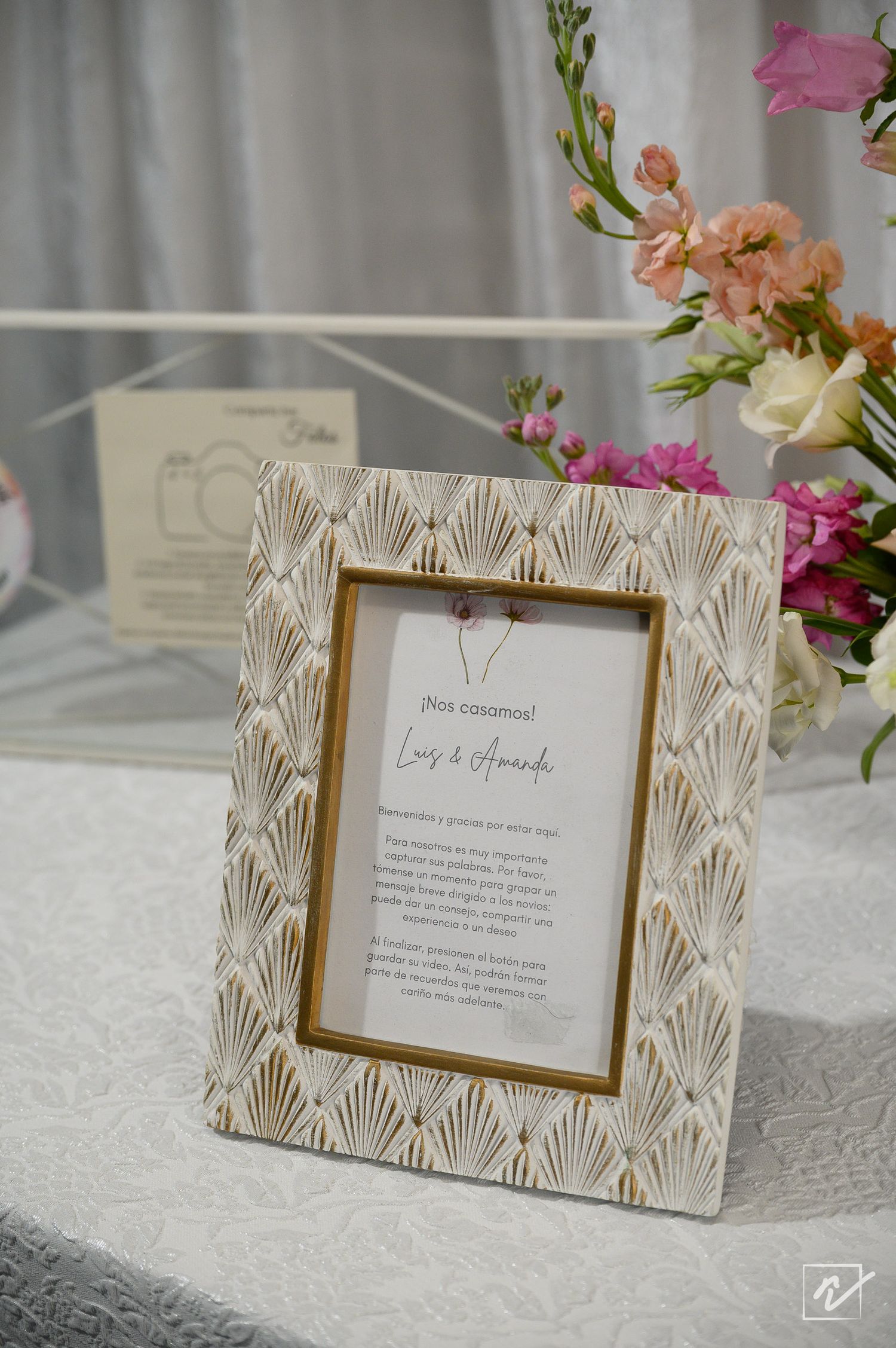Wedding thank-you card in an ornate gold art deco picture frame on a white tablecloth beside pink and peach floral arrangements.