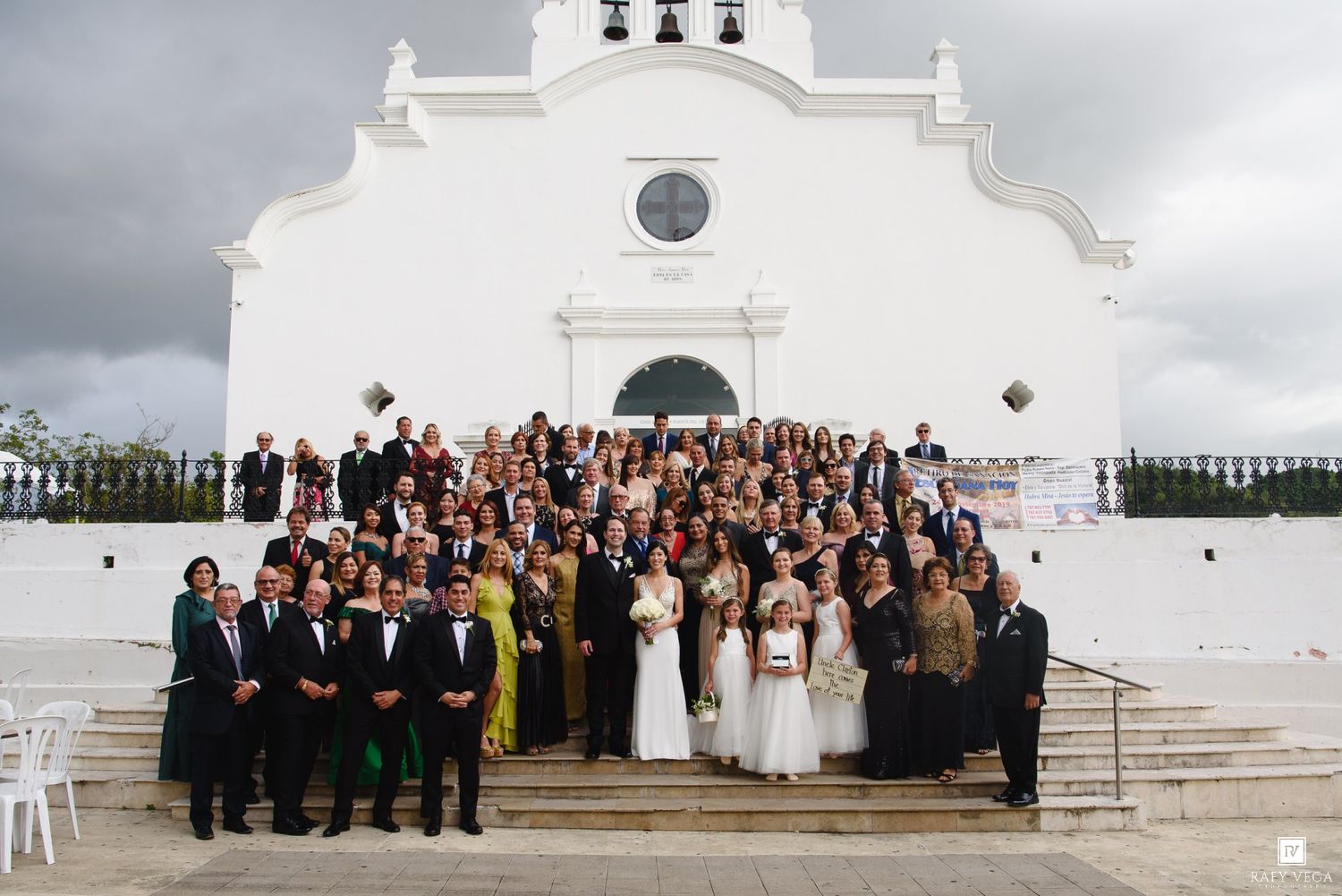 Large wedding party group photo on steps of a white church with guests in formal attire.
