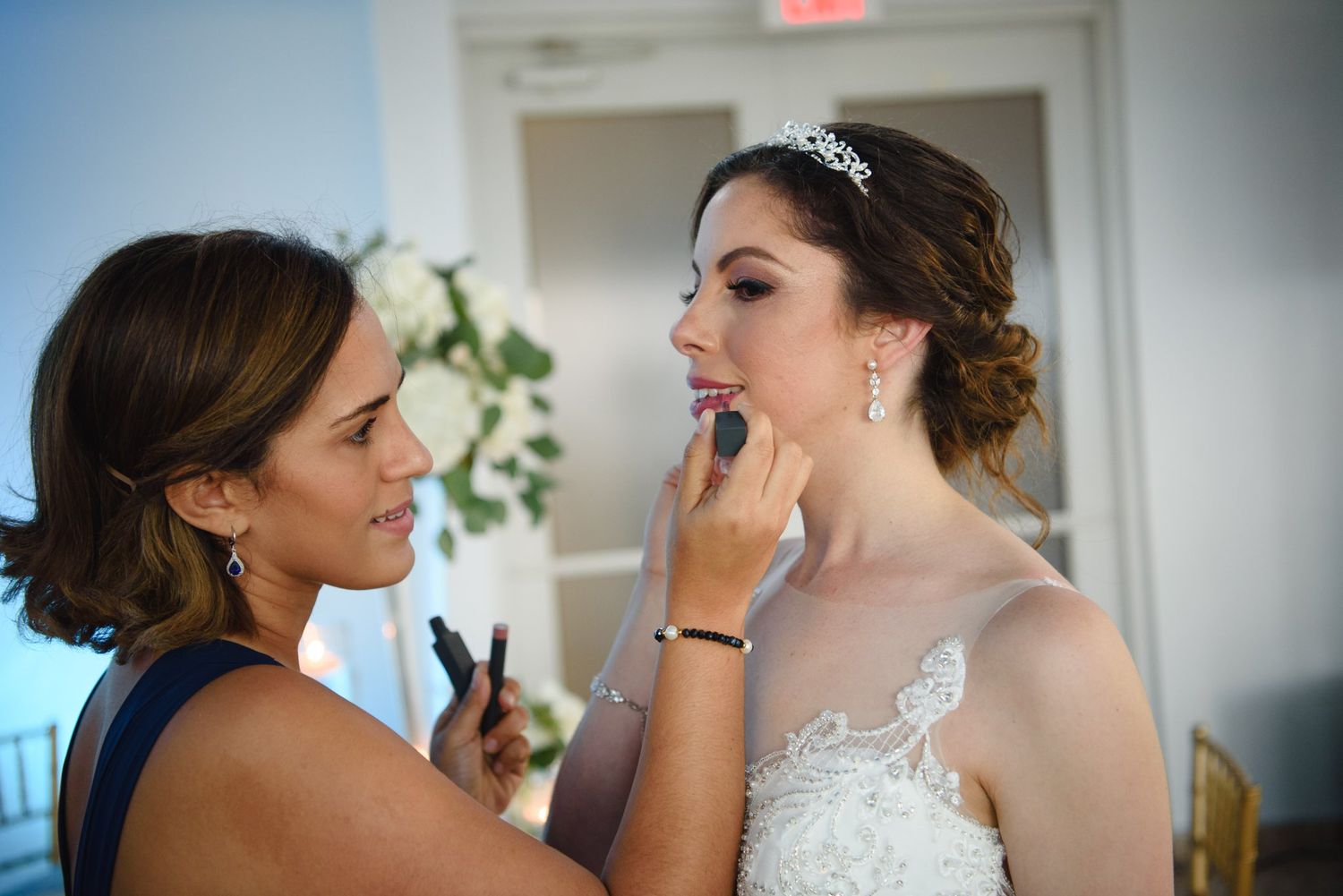 Makeup artist applying lipstick to a bride in a lace wedding dress and tiara before her wedding ceremony.