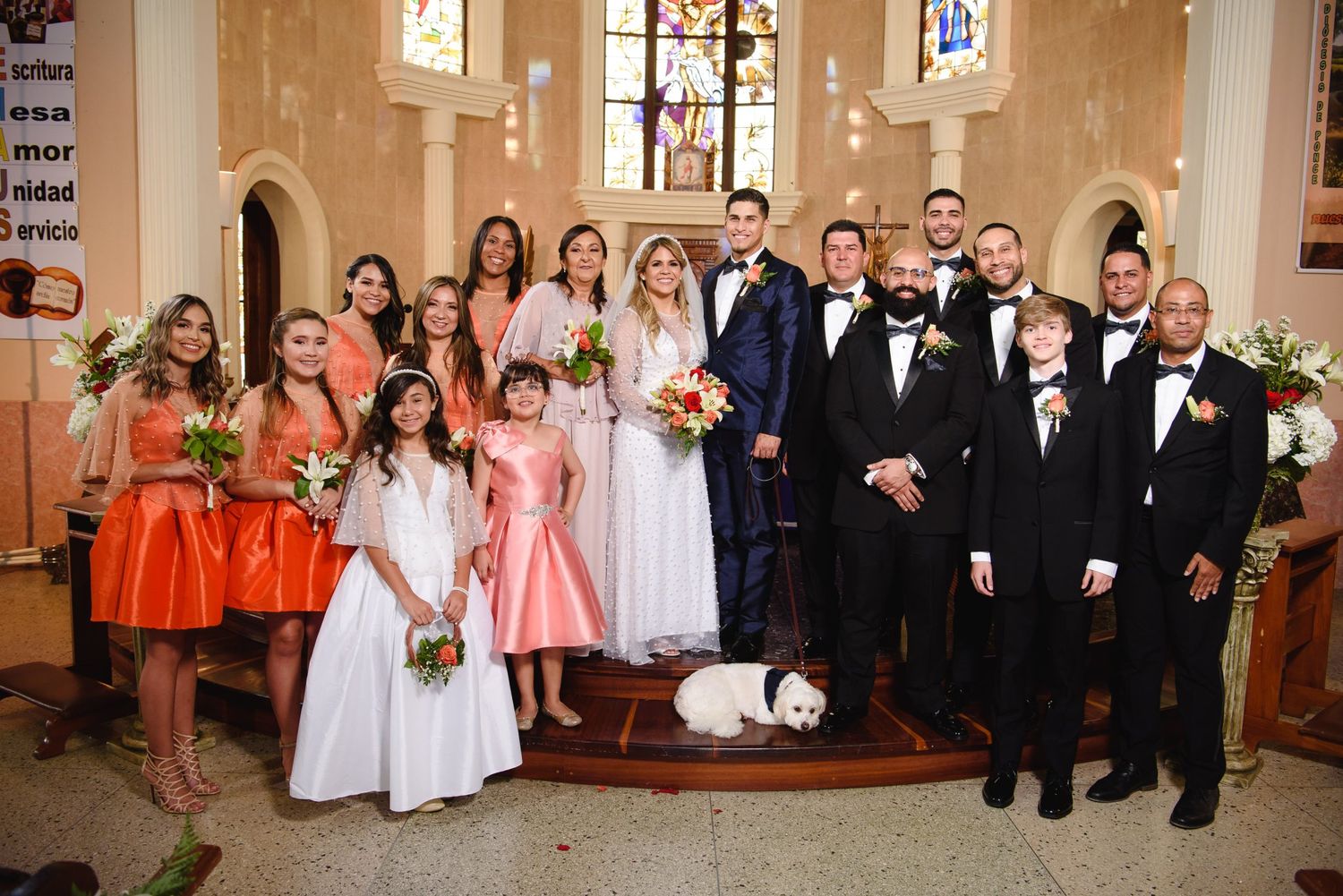 Wedding party poses in a church with bridesmaids in orange dresses, groomsmen in suits, and bride and groom at center.