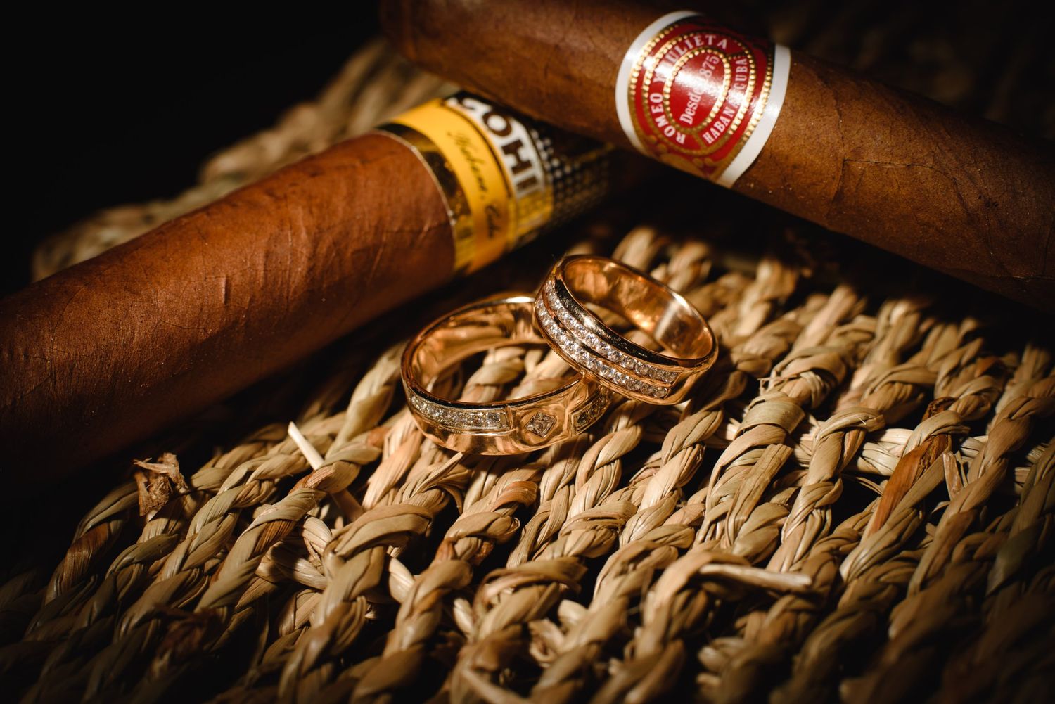 Two premium cigars and gold wedding rings resting on woven straw surface in dramatic dark lighting.