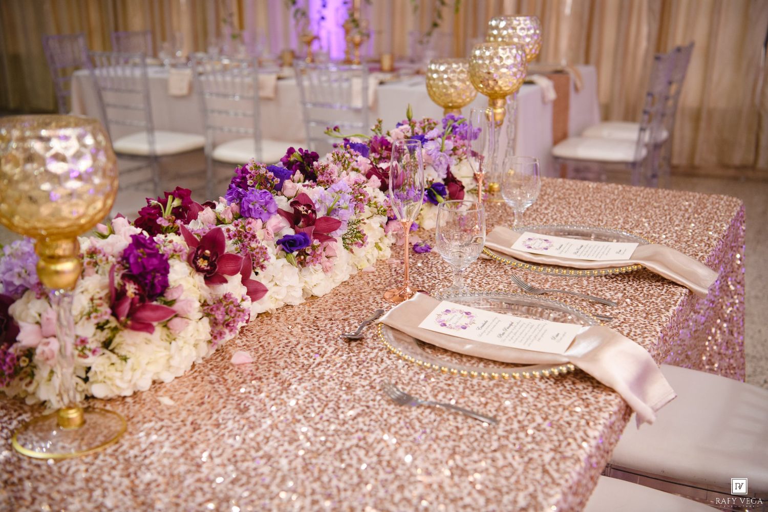 Elegant wedding reception table with rose gold sequin tablecloth, purple floral centerpiece, and gold candle holders.