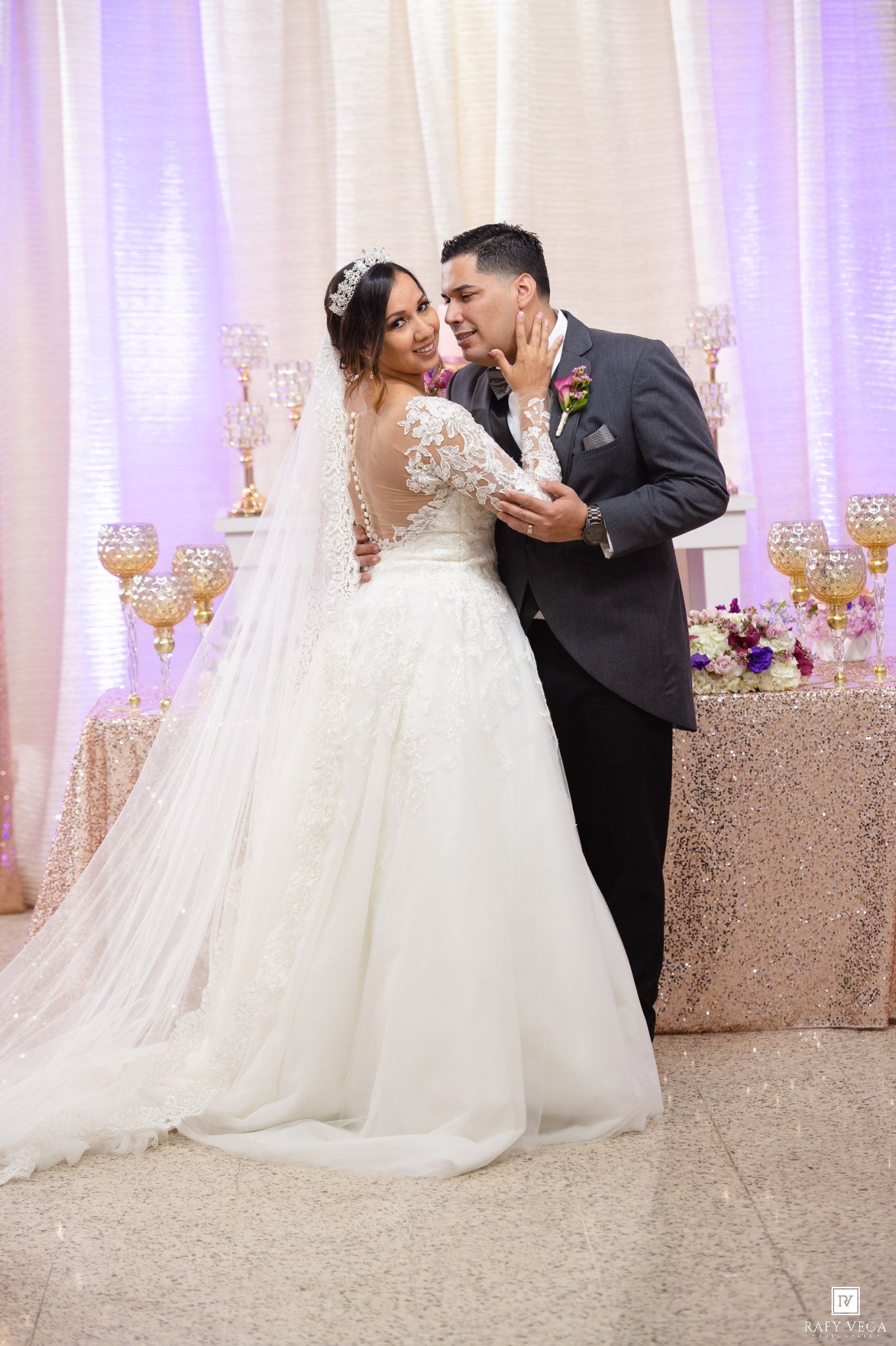 Bride in white lace gown and veil shares romantic moment with groom in gray suit at elegant wedding reception.