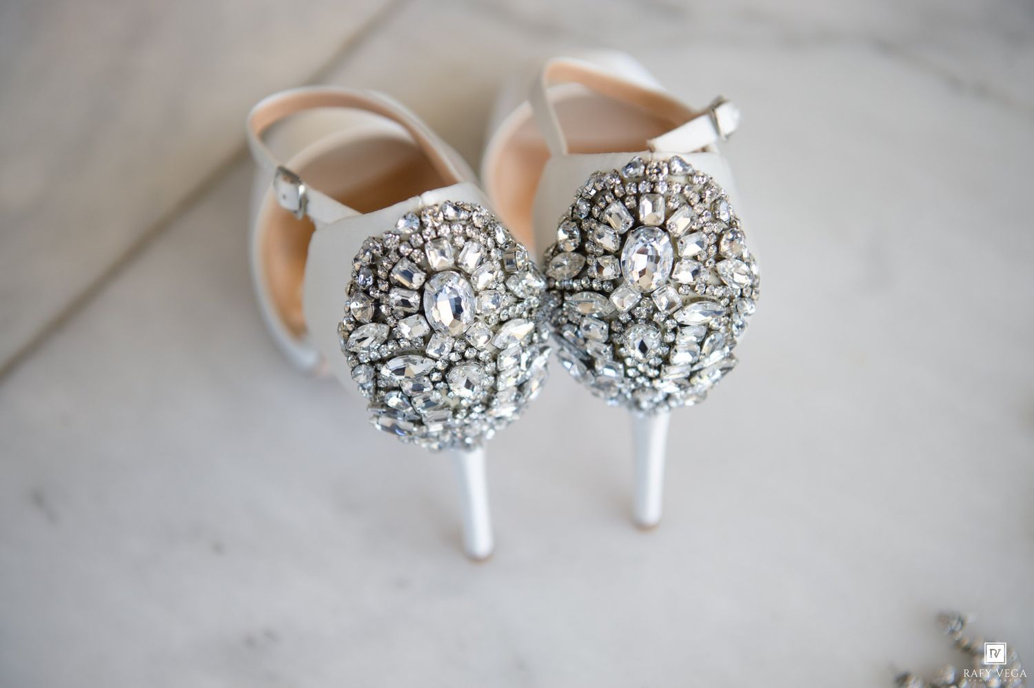 Elegant bridal heels adorned with sparkling crystal rhinestone embellishments on white satin pumps displayed on marble surface.