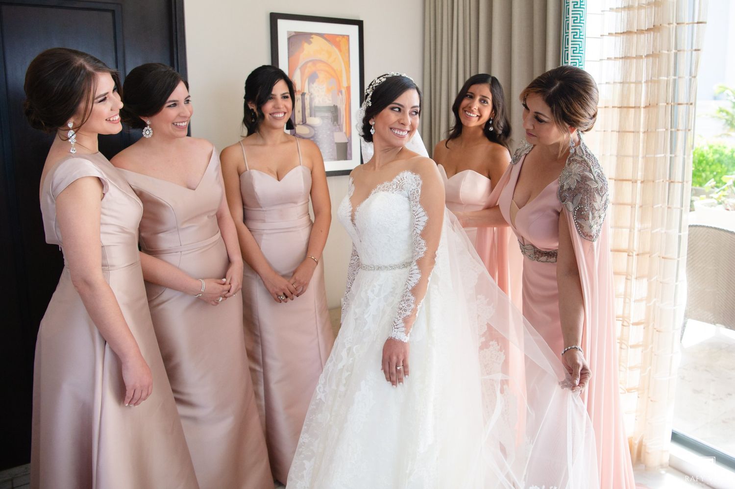 Bride in white lace gown smiles with five bridesmaids in blush pink dresses during wedding preparations.