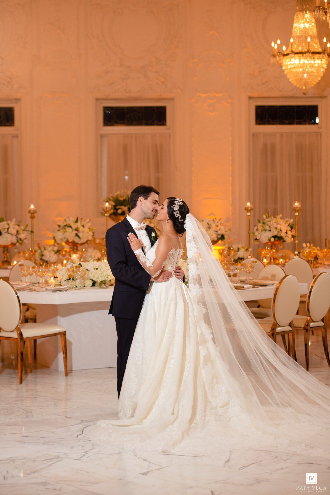 Bride and groom share a romantic kiss in an elegant ballroom with golden lighting, floral centerpieces, and a cathedral veil.