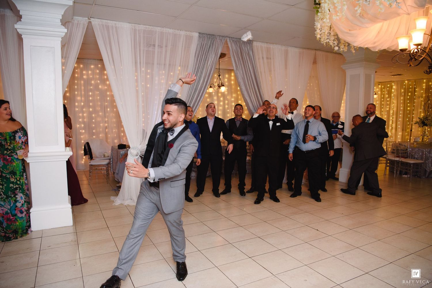 Groom in gray suit tosses garter to single men on wedding reception dance floor.