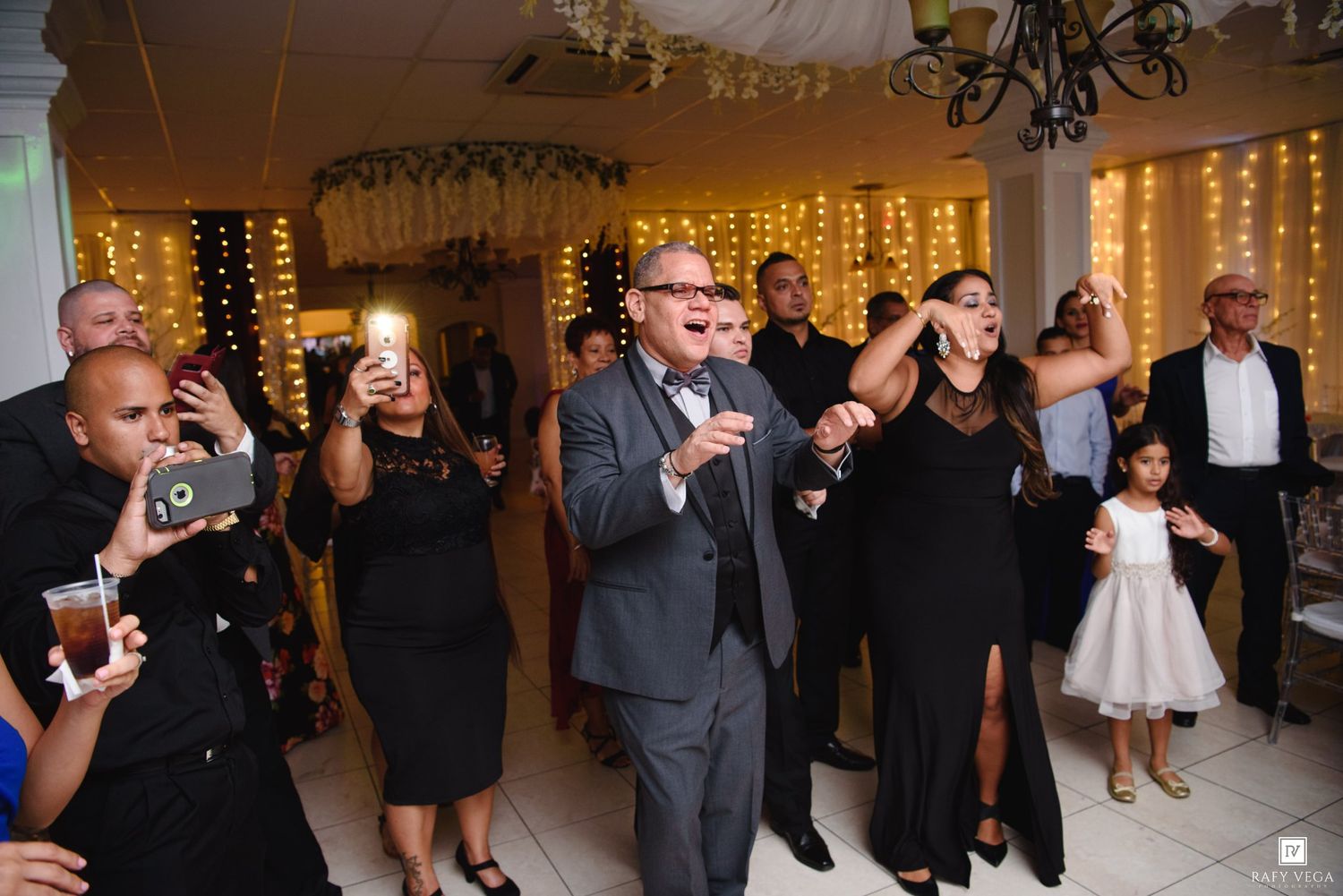 Guests celebrate at a wedding reception with fairy lights, dancing and taking photos in an elegant venue.