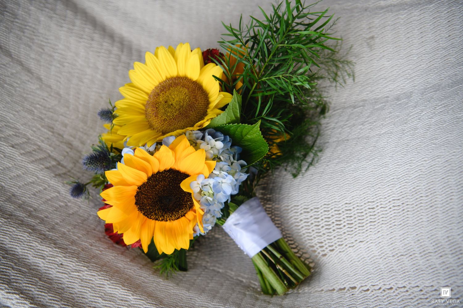 A bridal bouquet featuring two bright yellow sunflowers with blue hydrangeas and green ferns wrapped in white ribbon.
