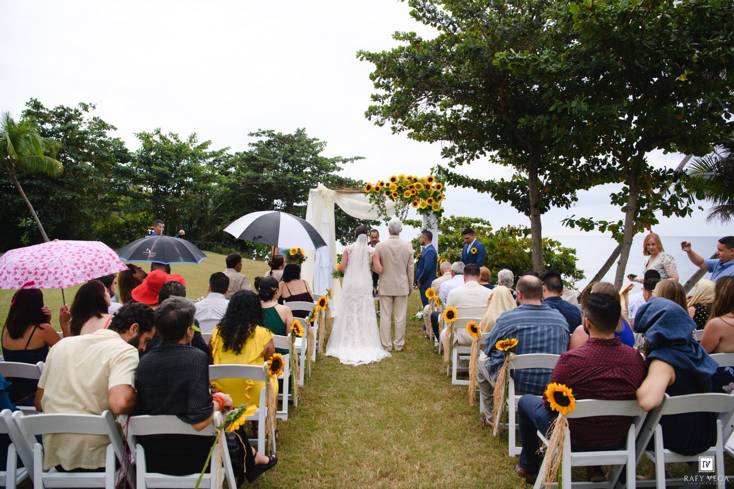 Outdoor wedding ceremony on green lawn with guests seated in white chairs under trees, bride and groom at sunflower arch.