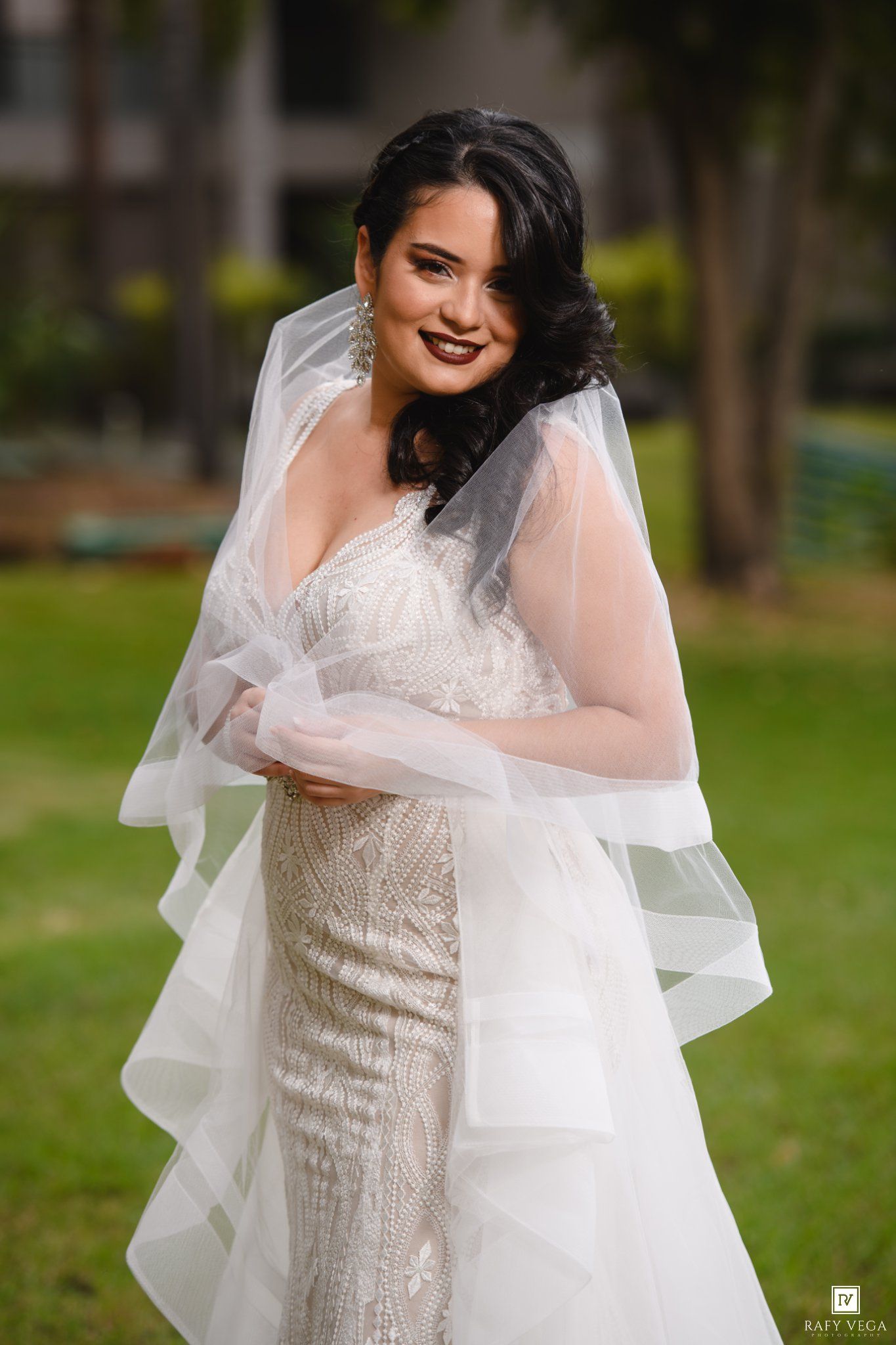 Smiling bride in beaded white gown with layered veil poses outdoors on sunny day.