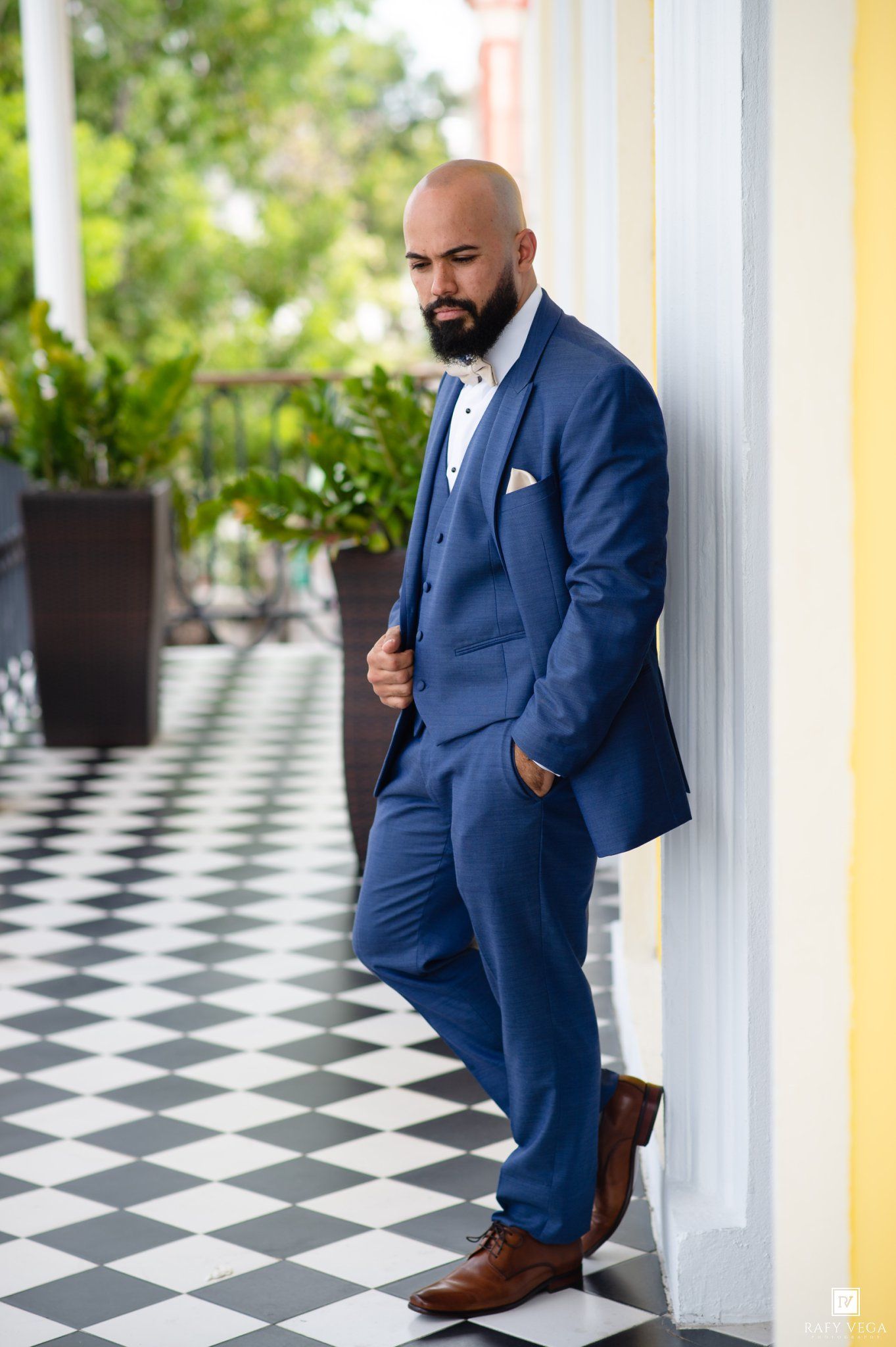 Bald bearded groom in blue three-piece suit with bow tie leaning against yellow column on checkered floor.