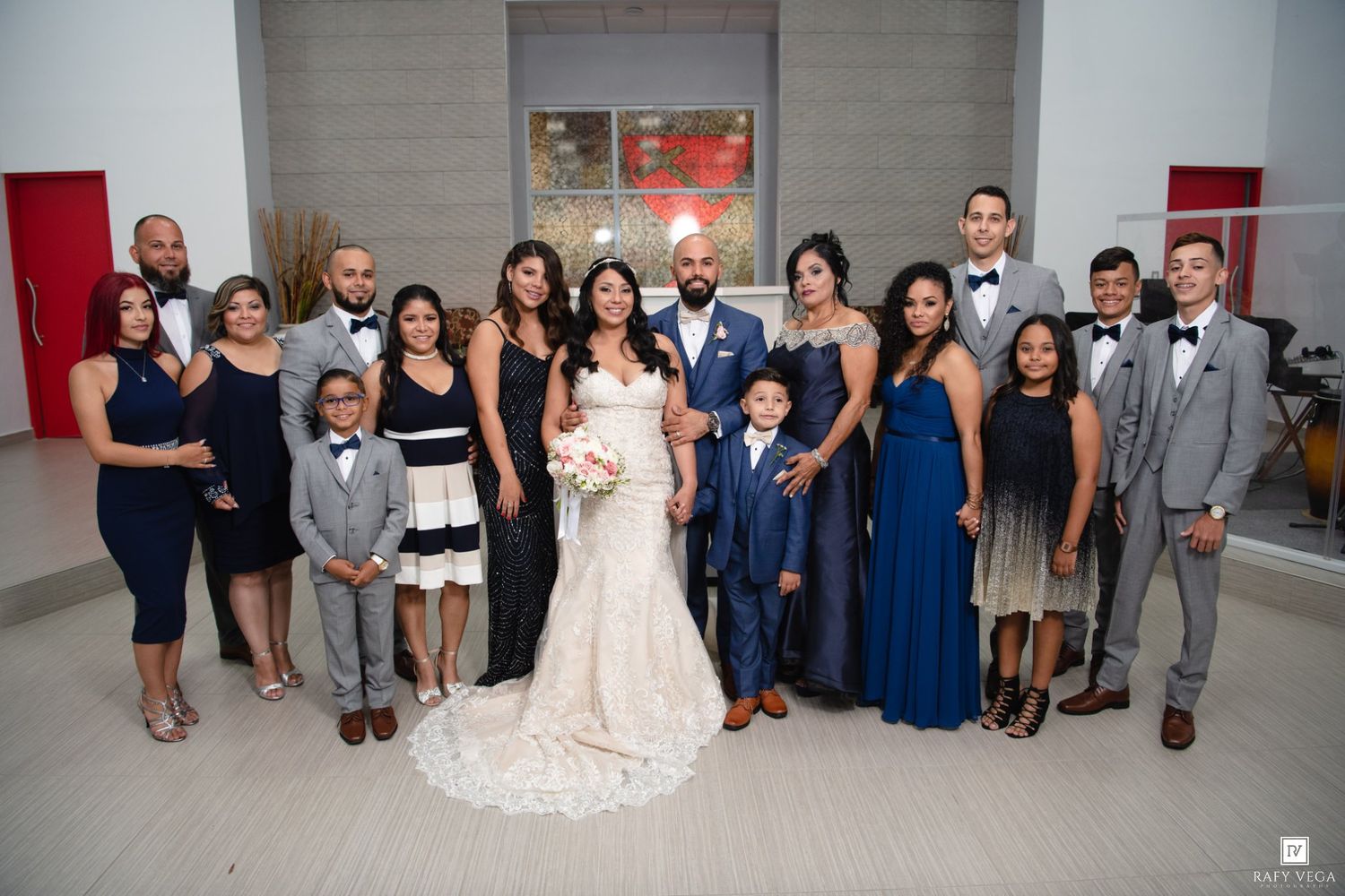 Wedding party poses together in formal attire, bride in white gown with bouquet, groom in blue suit, indoors.