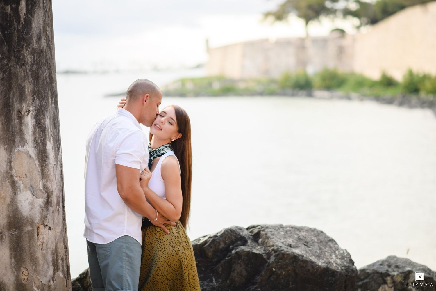 Romantic couple embracing near waterfront stone wall, woman in gold skirt smiling up at man in white shirt.